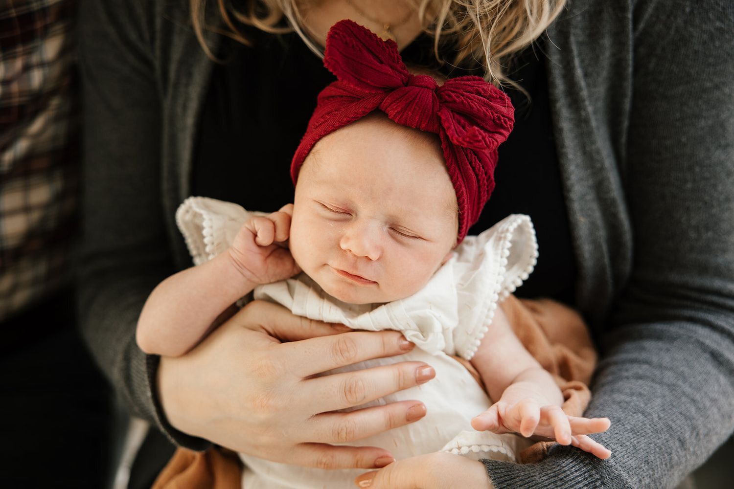 Baby wearing burgundy bow lies in white outfit while being cradled.