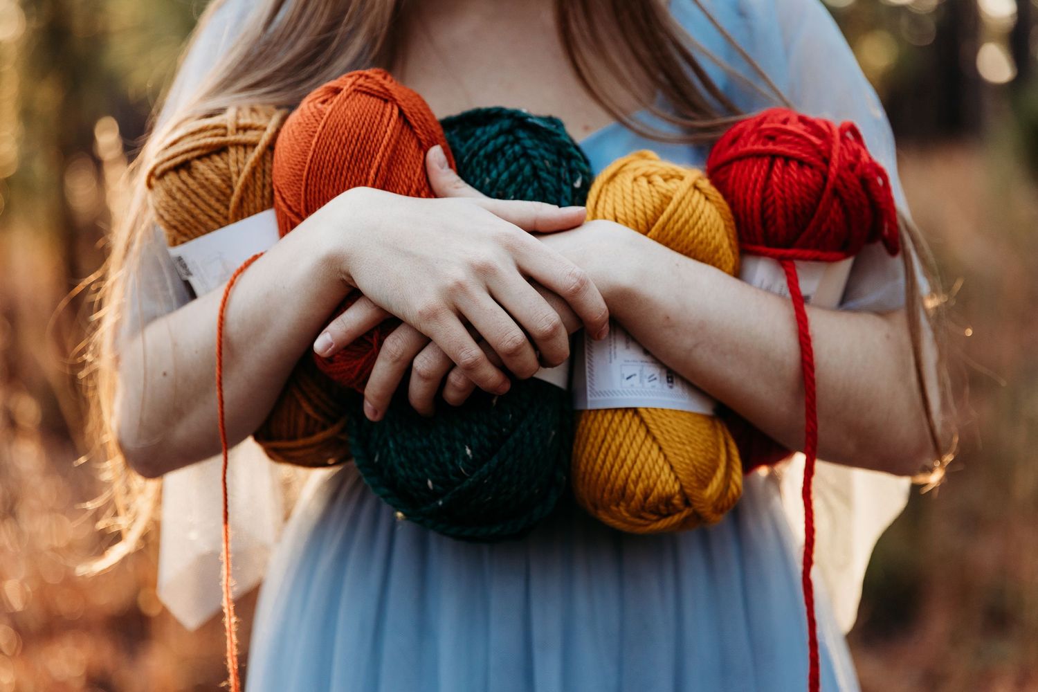 Person in a blue dress holds colorful yarn balls against a warm sunset background.