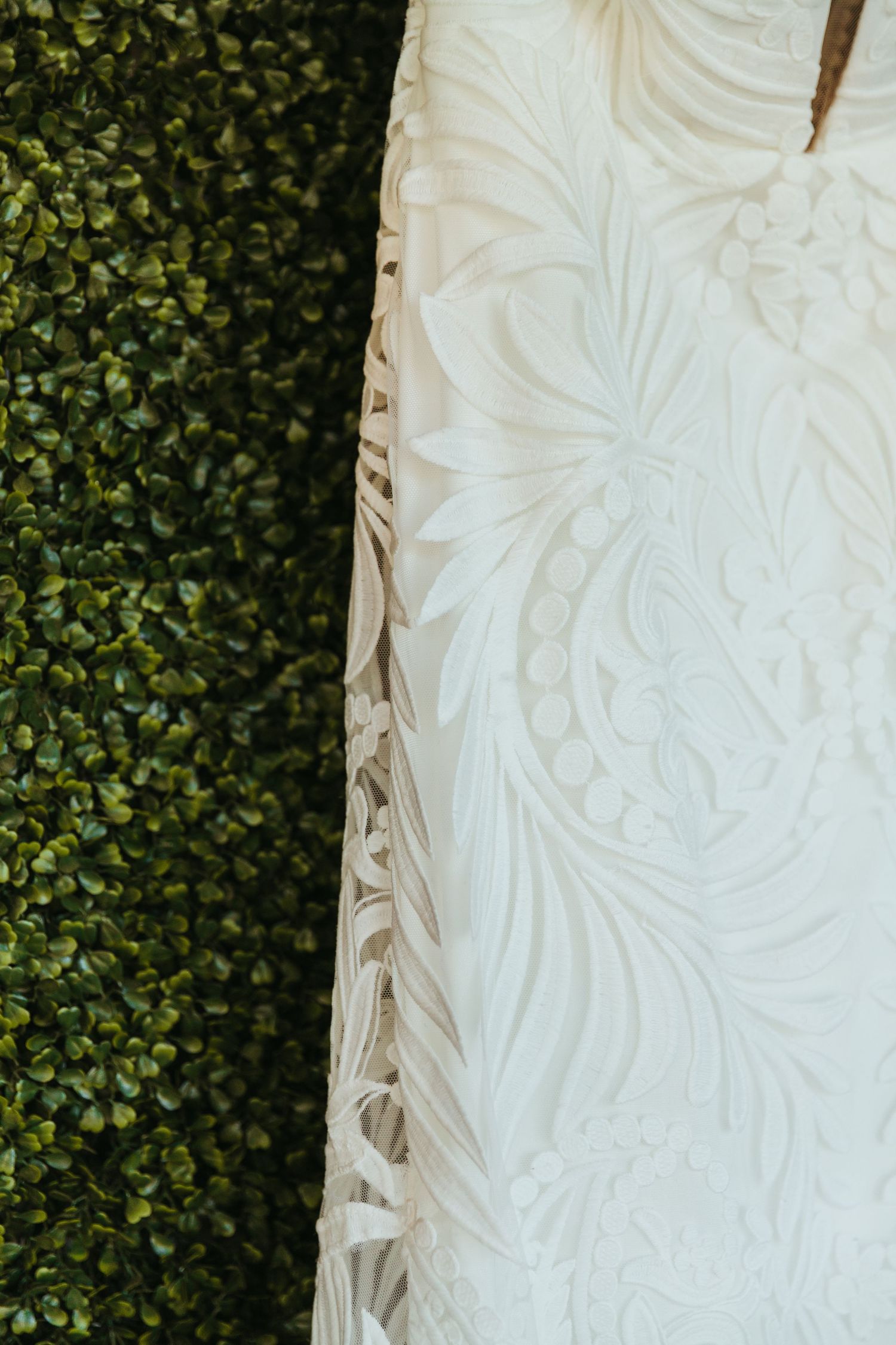 Close-up detail of an intricate white lace wedding dress pattern against a green hedge background.