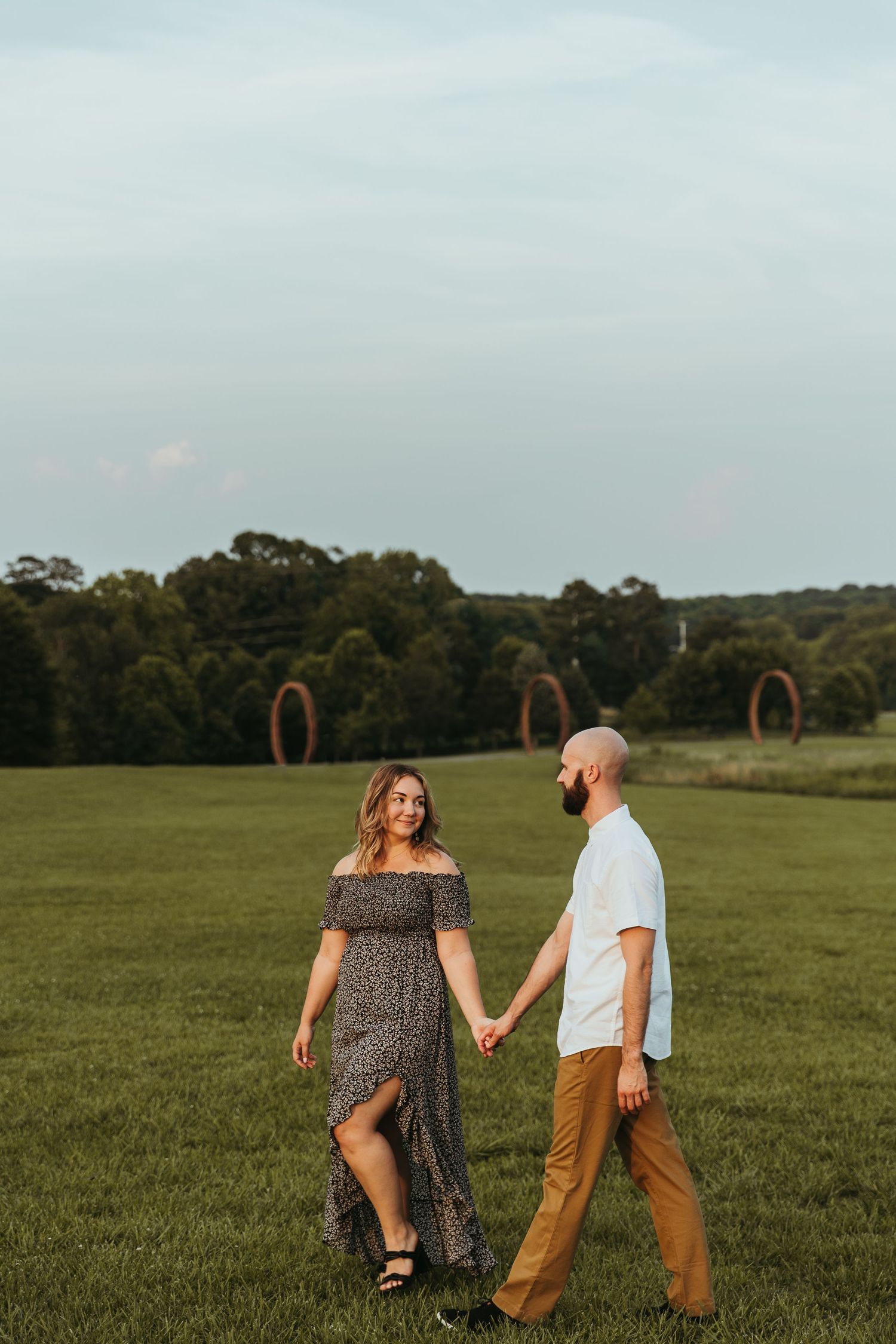 A couple holds hands while walking together in a grassy field on an overcast day.