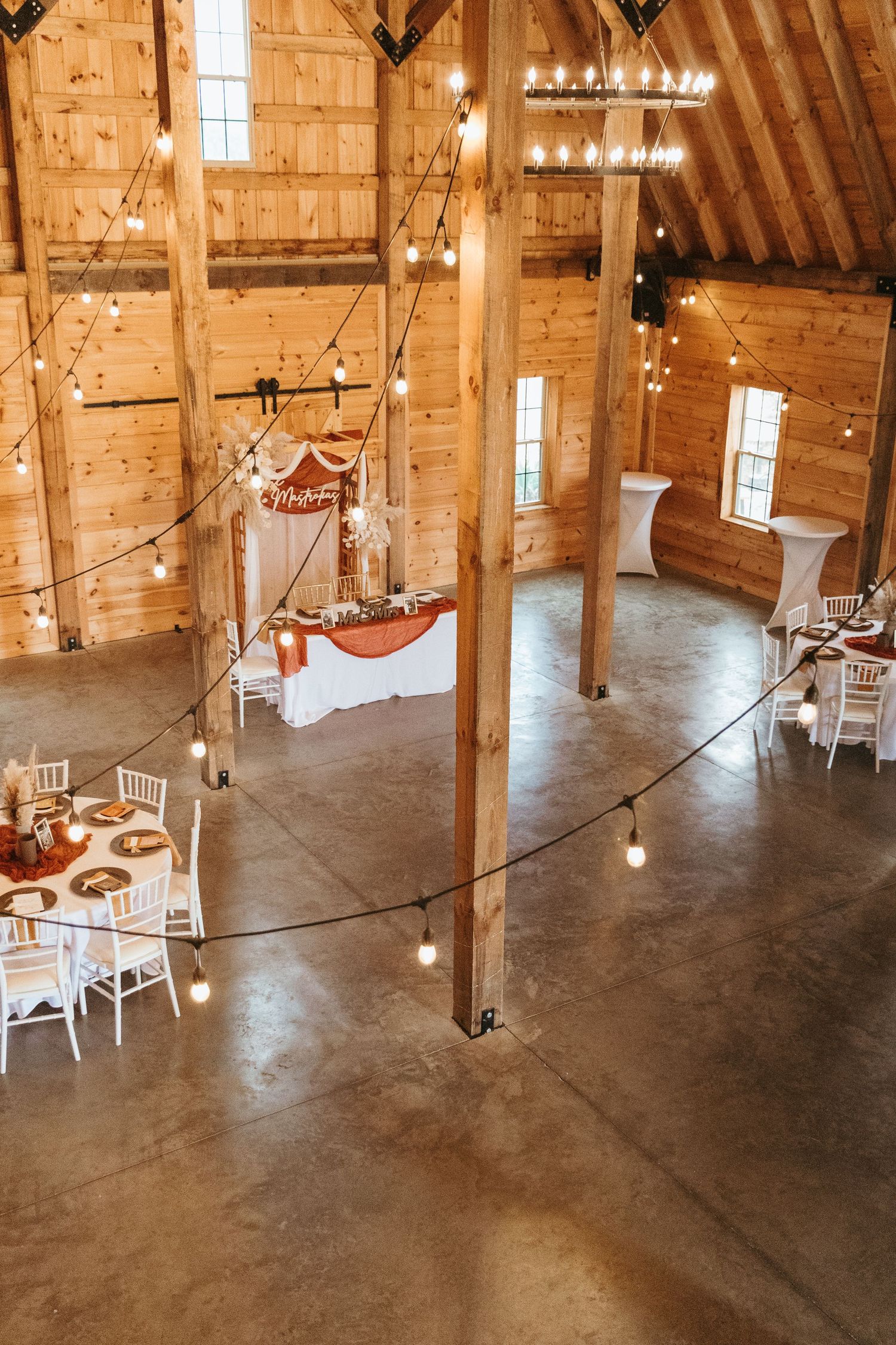 Interior of a rustic wooden barn venue with string lights, chandeliers and white tables set up for an elegant event.