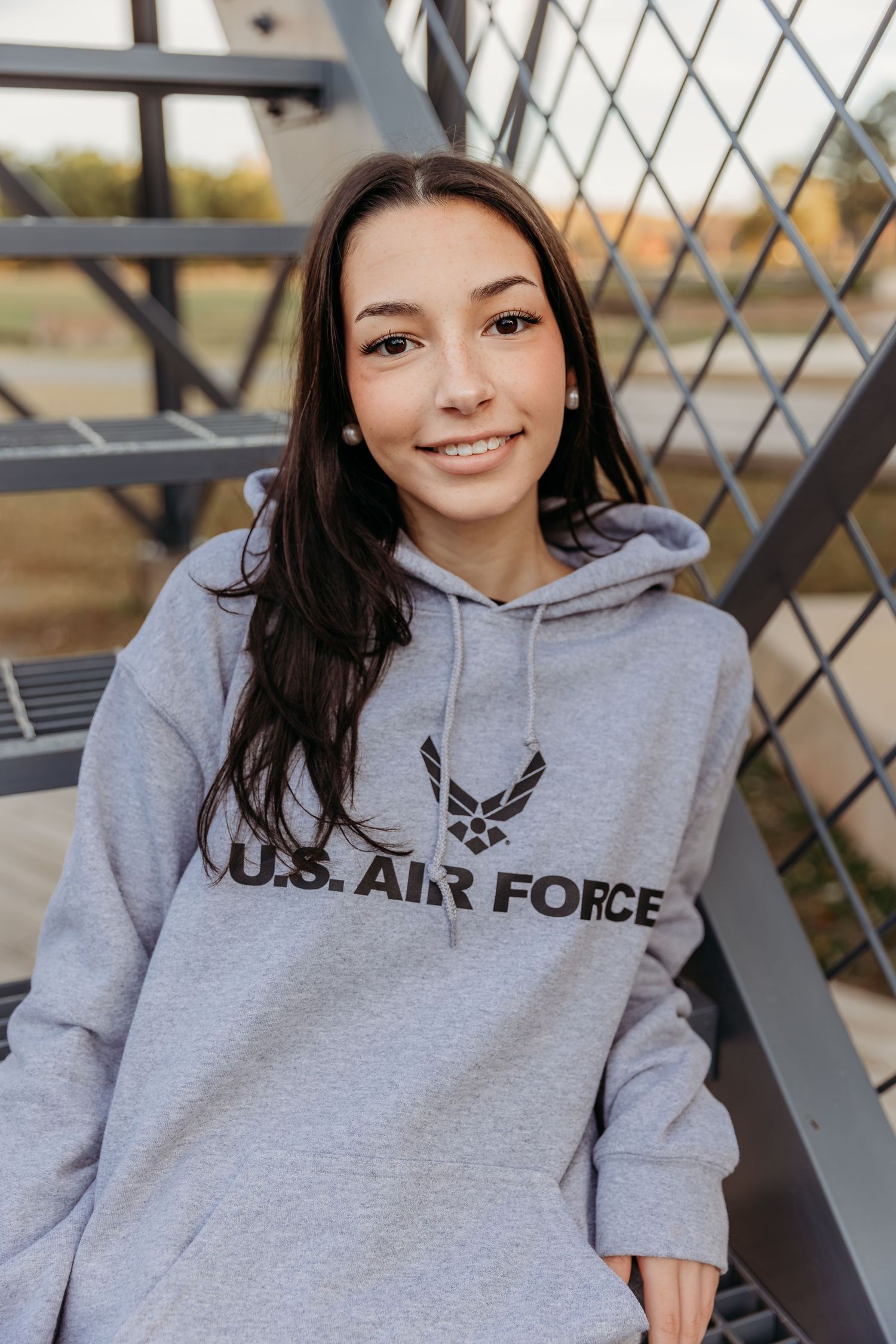 A person wearing a gray U.S. Air Force hoodie sweatshirt poses against a metal fence background.