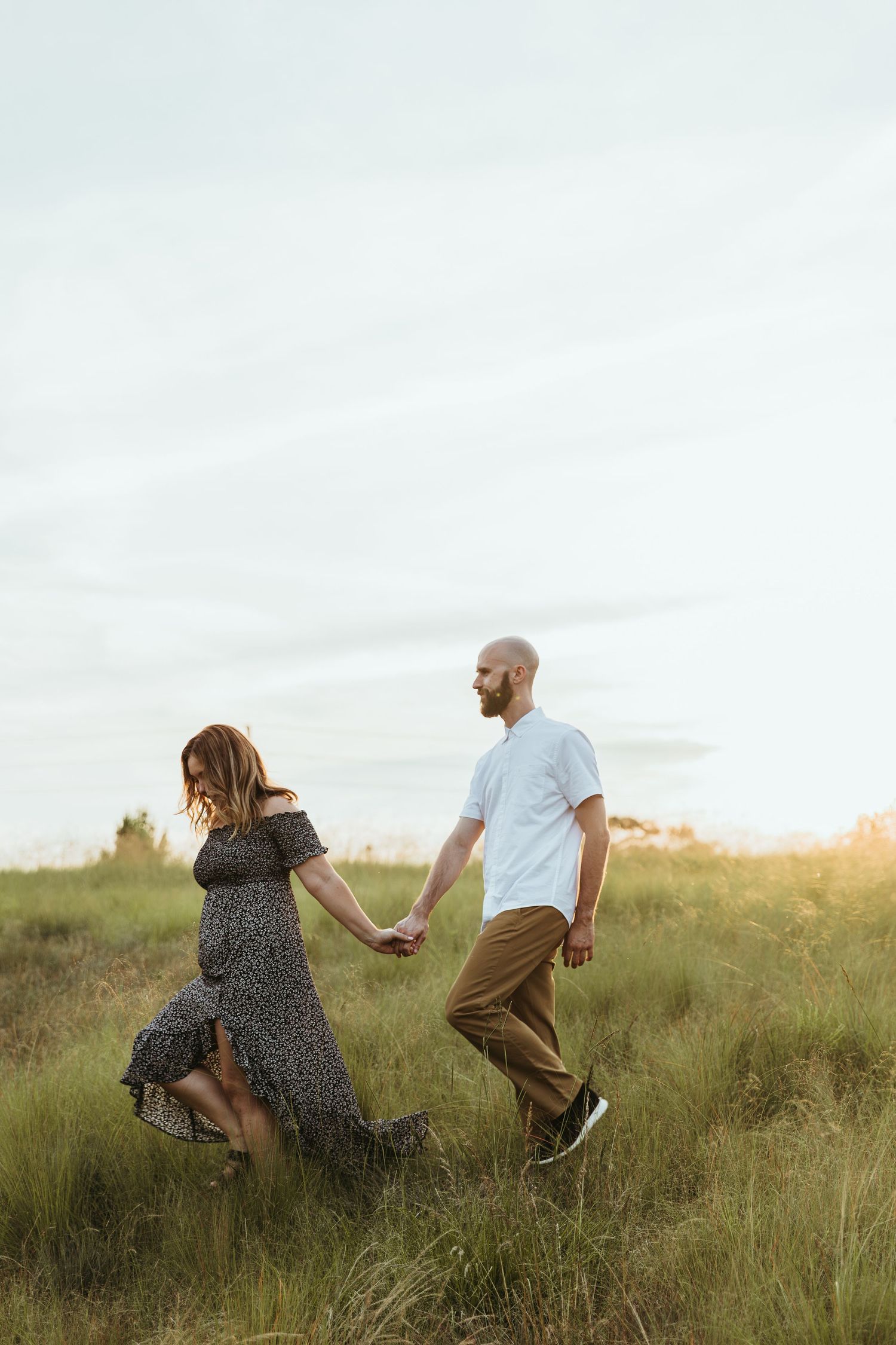 A couple playfully runs and jumps through a grassy meadow at sunset creating a joyful silhouette against the sky.