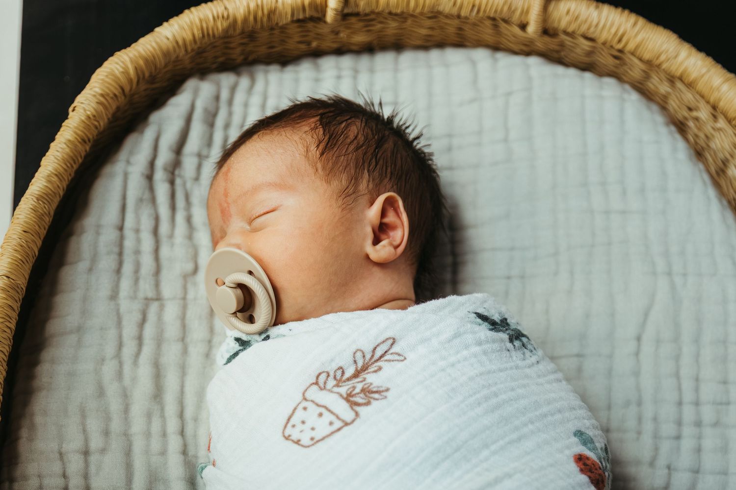 A sleeping newborn swaddled in white fabric with a pacifier rests peacefully in a woven bassinet.