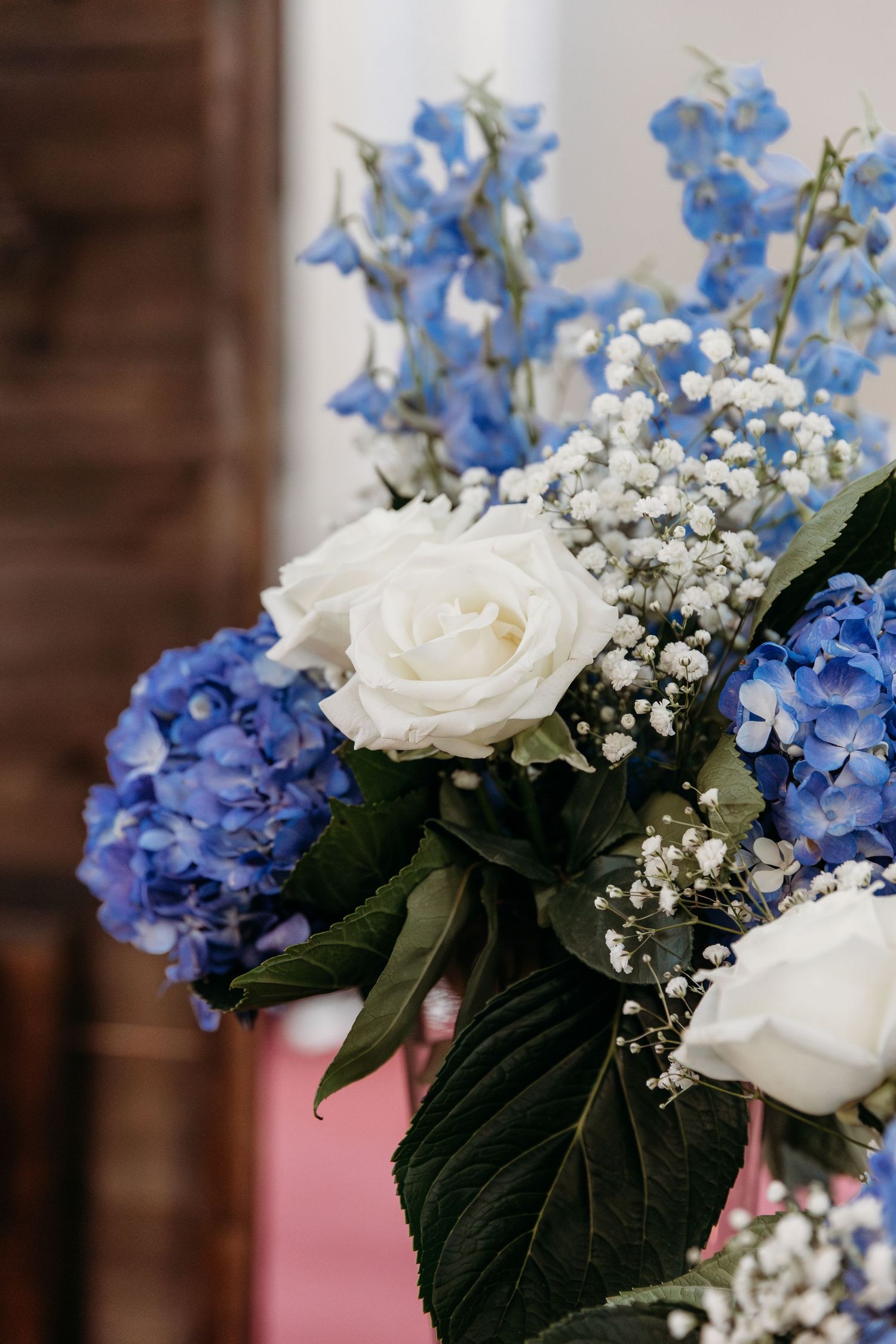 Close-up of white roses and blue hydrangeas with baby's breath flowers arranged against a blurred brick wall background.