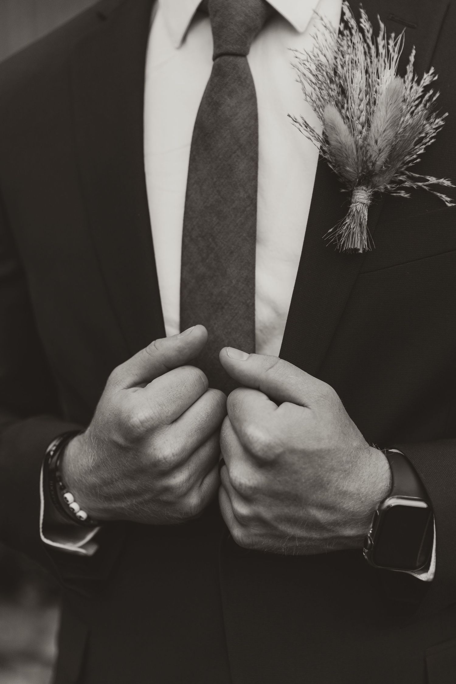 Black and white close-up image showing hands adjusting a dark tie against a white dress shirt and suit jacket.