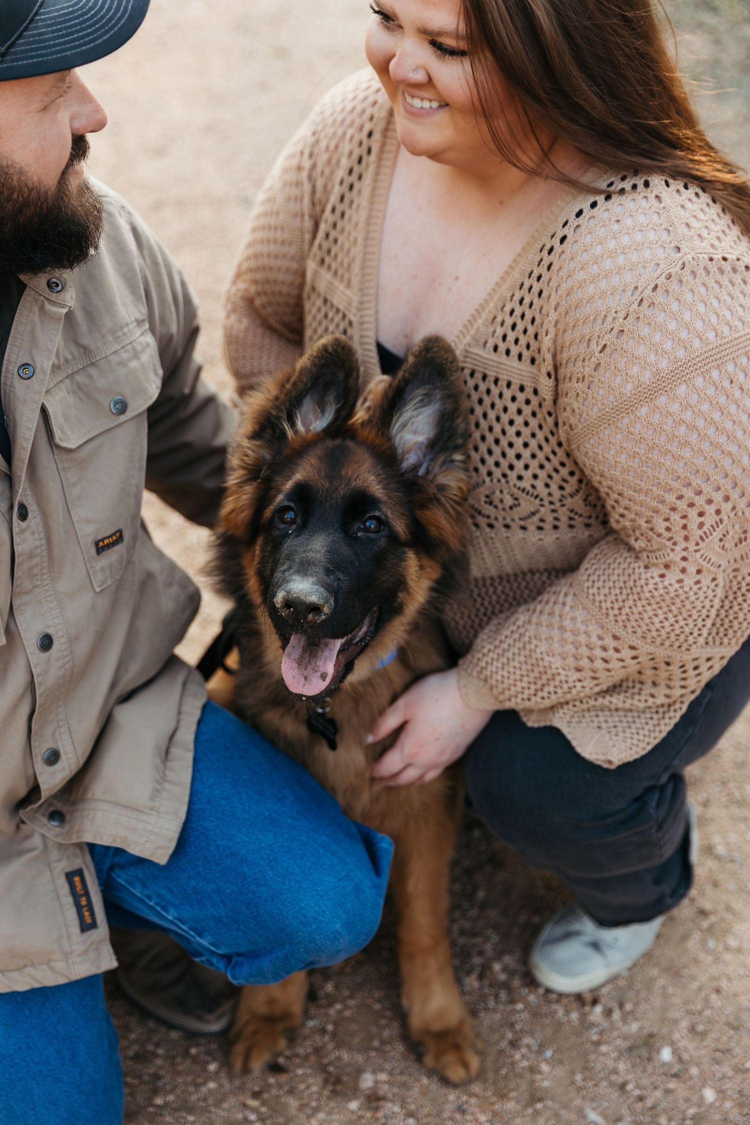 A happy German Shepherd puppy sits between two people wearing casual clothing on a dirt path.