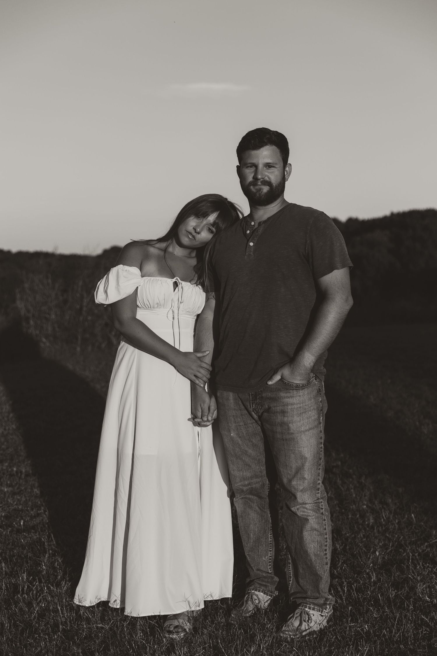 A couple stands together in a grassy field at dusk, with the person in white leaning against their partner.