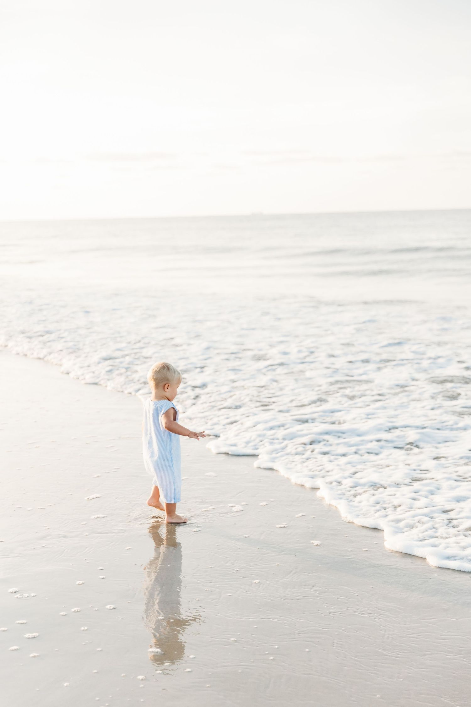 A toddler in a light blue outfit walks along the shoreline as gentle waves roll onto a sandy beach.