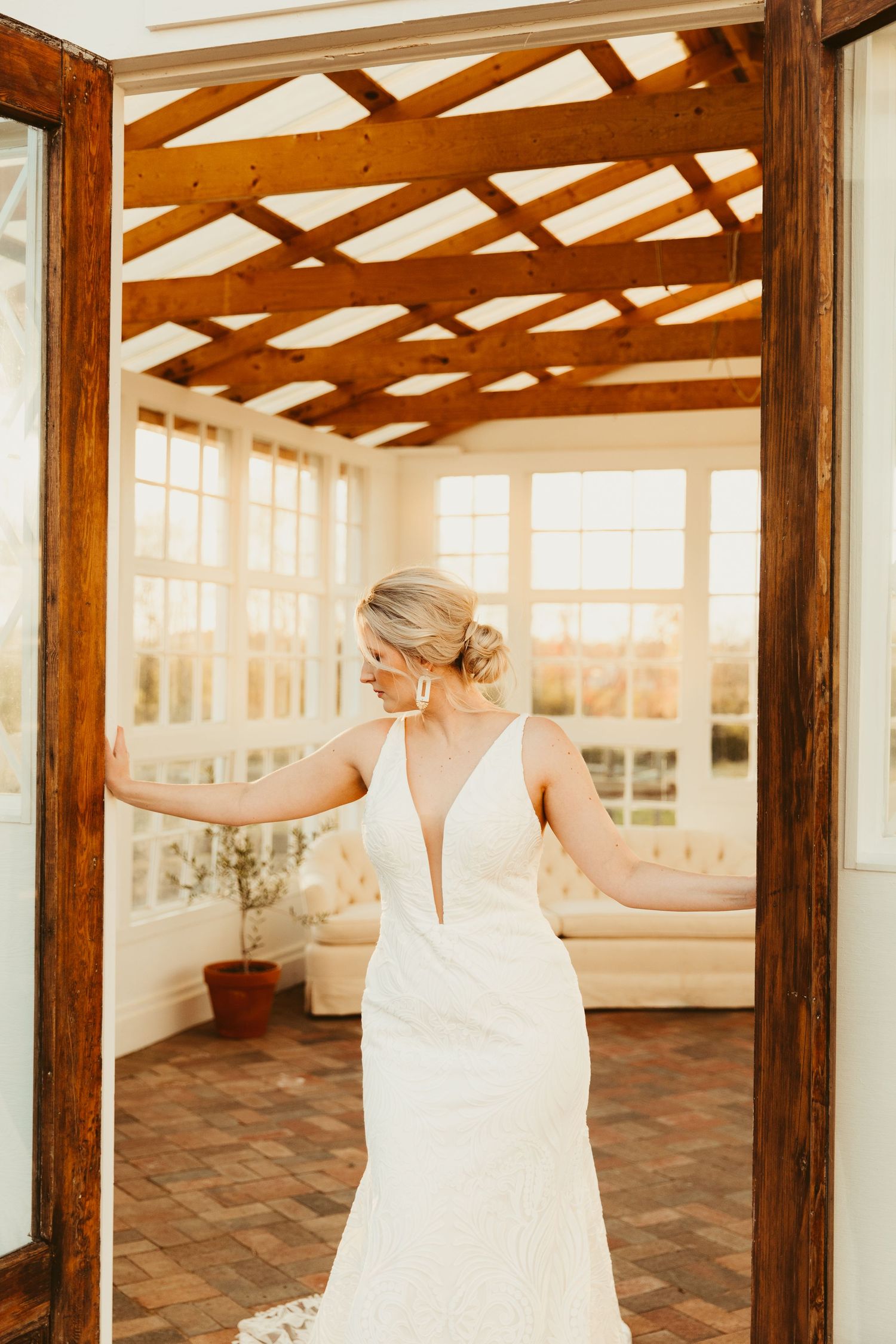 A person in a white dress stands in a rustic wooden doorway with large windows and natural light streaming in.