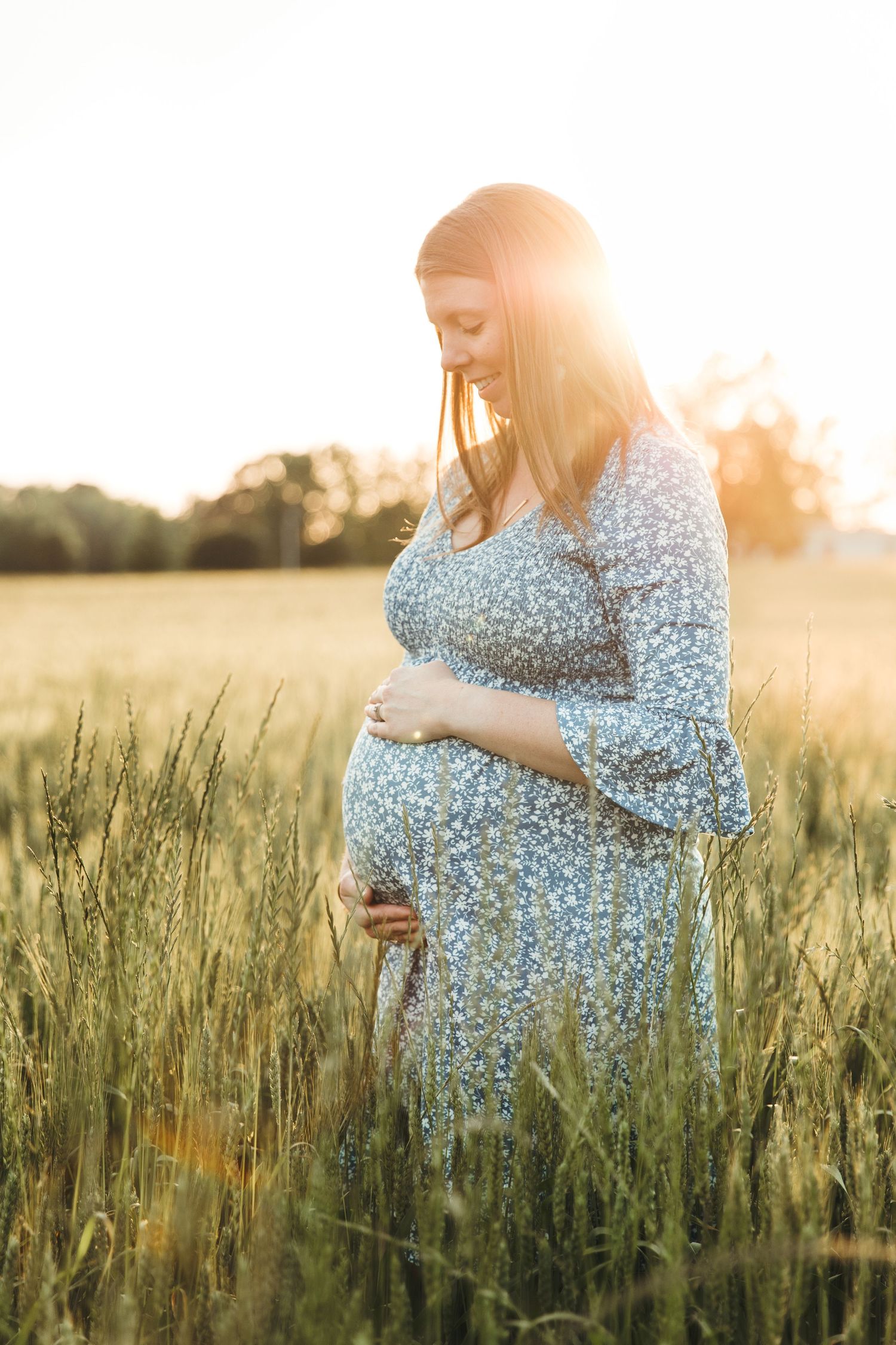 Expectant woman in floral dress stands in golden wheat field at sunset while cradling her belly.