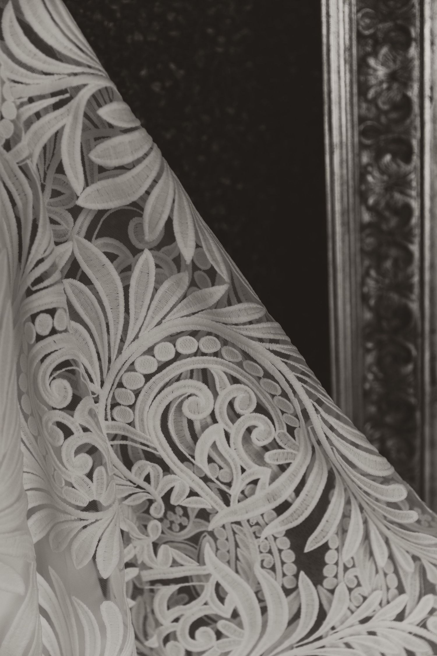 black and Close-up detail of an intricate white lace wedding dress pattern against a green hedge background.