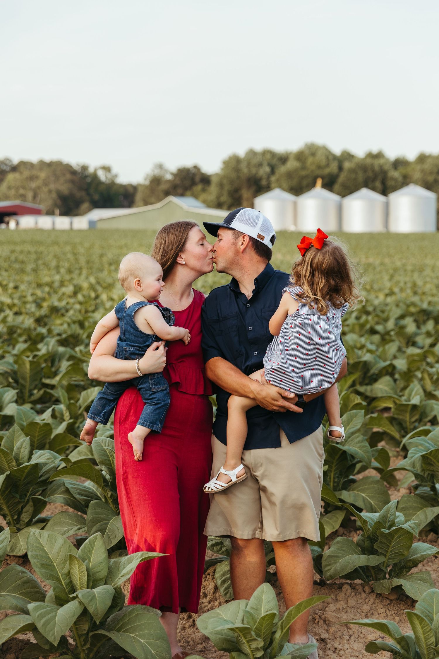 A family shares a tender moment together while standing in a lush green tobacco field with grain silos in the background.