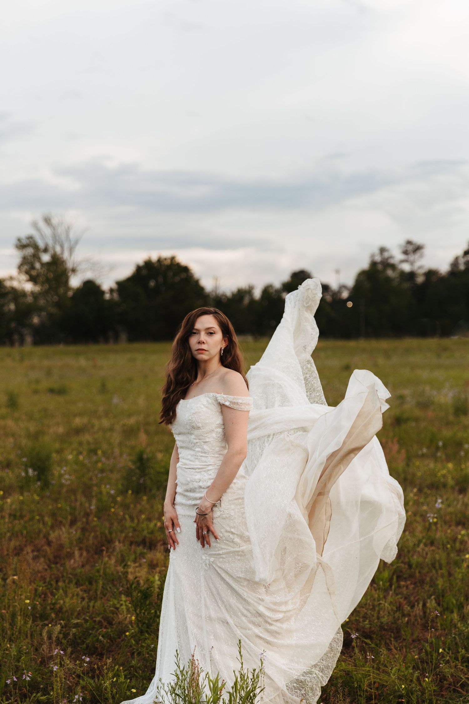 A flowing white wedding dress billows gracefully in the breeze as someone poses in a rustic field at sunset.