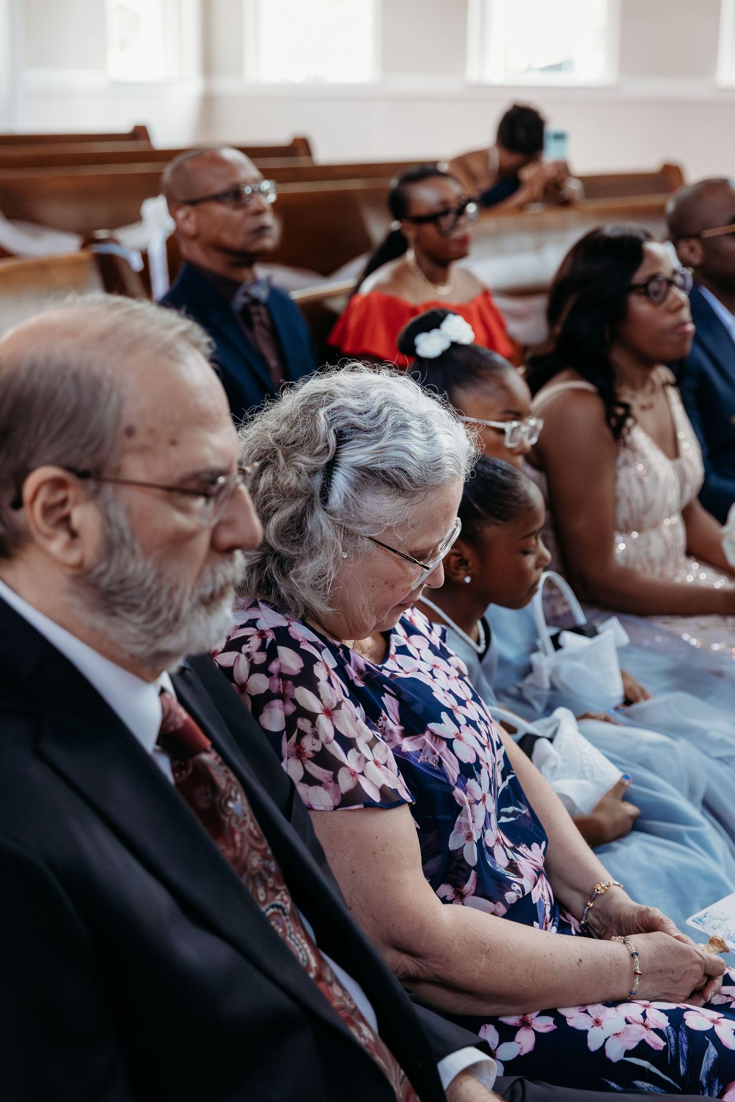 A diverse congregation sits in wooden pews during a church service wearing formal attire and glasses.