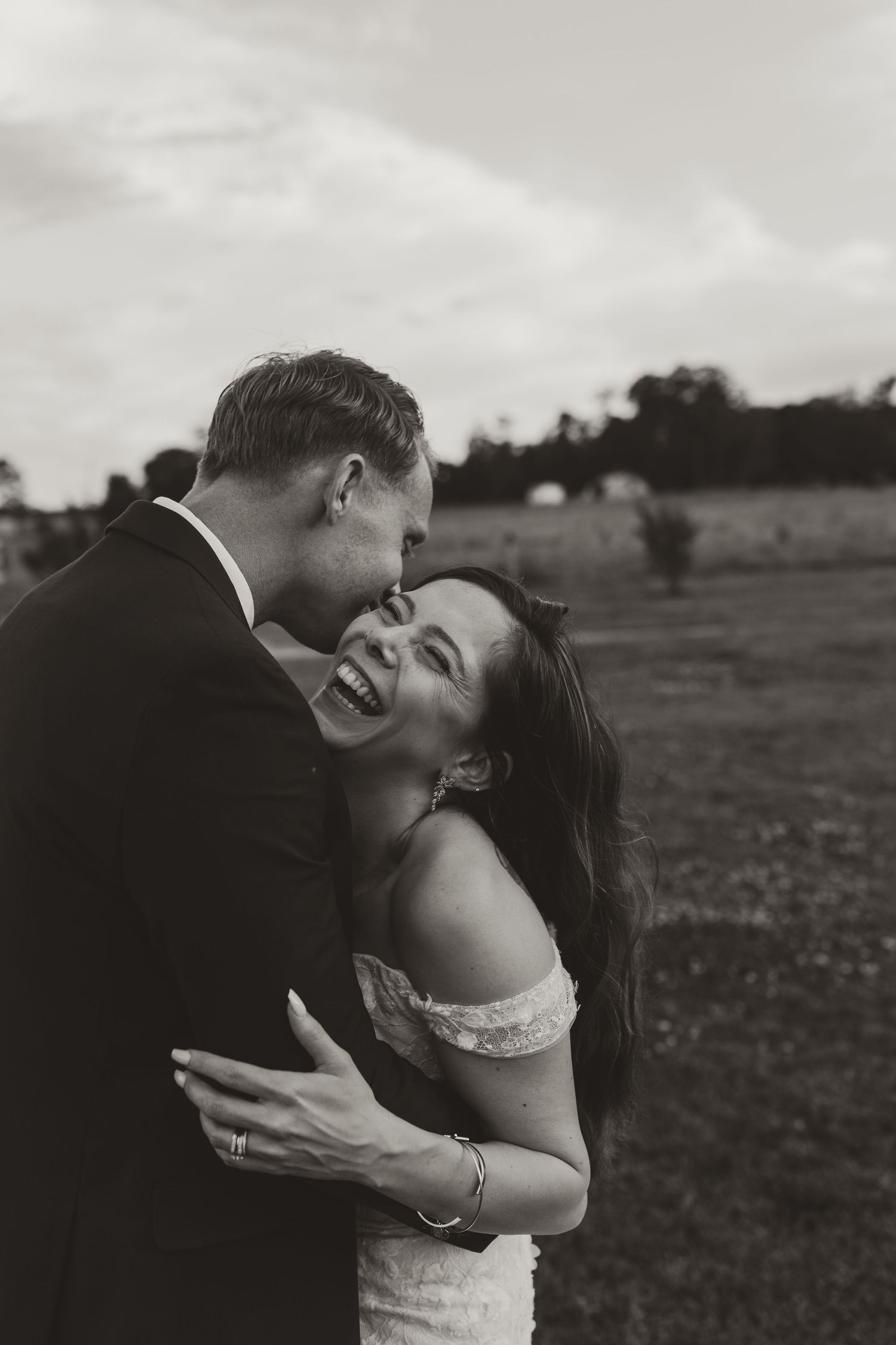Black and white romantic wedding photo of a couple sharing a joyful embrace in an outdoor field.