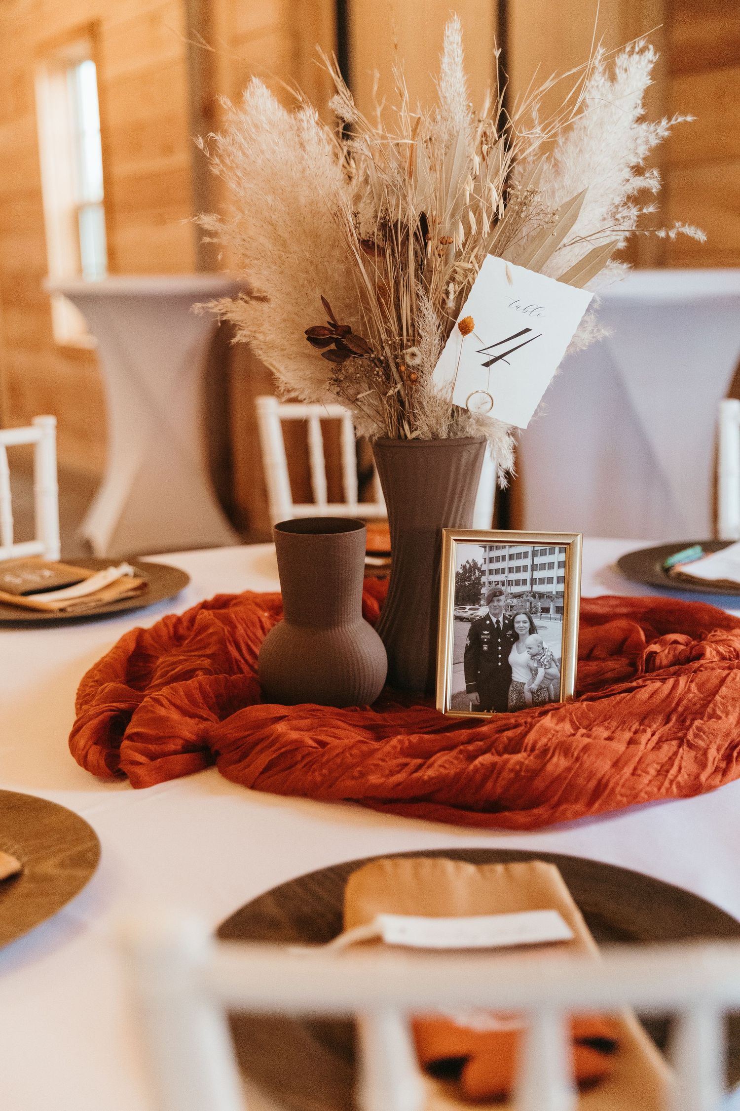 Wedding table centerpiece with pampas grass in brown vase, vintage photo frame, and burnt orange table runner on white tablecloth.
