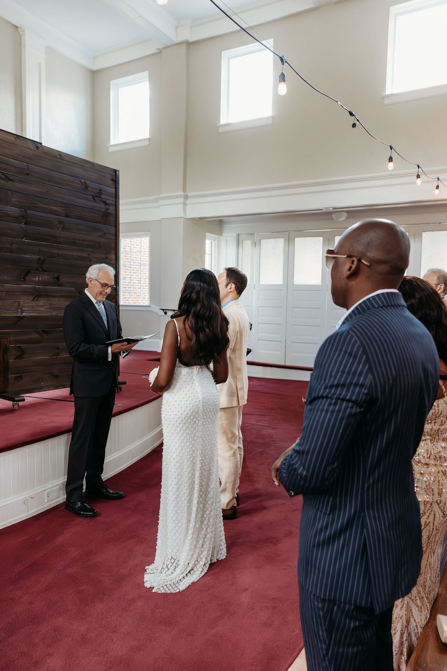 A couple in formal attire stands together in a church with white walls and red carpeting during a ceremony.