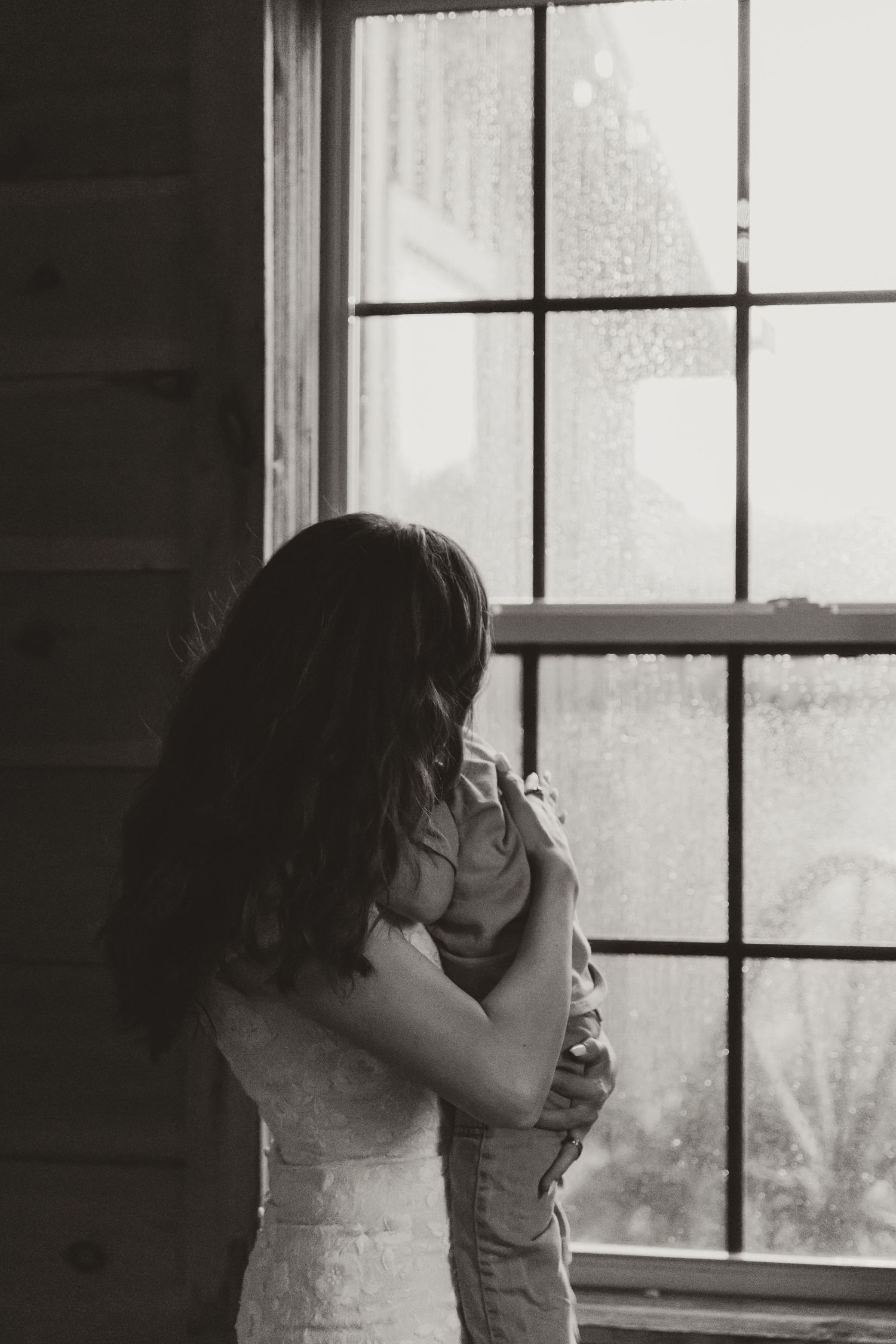 A mother in wedding dress holding her child by a window covered in rain drop