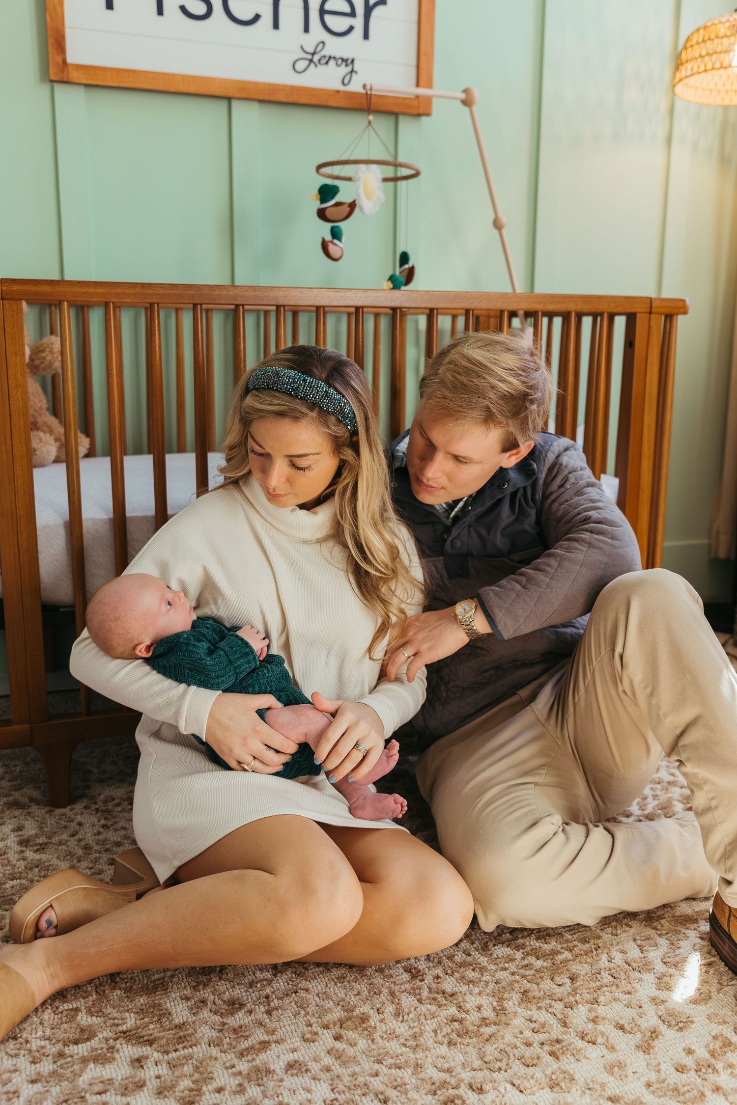 A couple sits together on a carpeted floor near a wooden crib in a nursery setting.