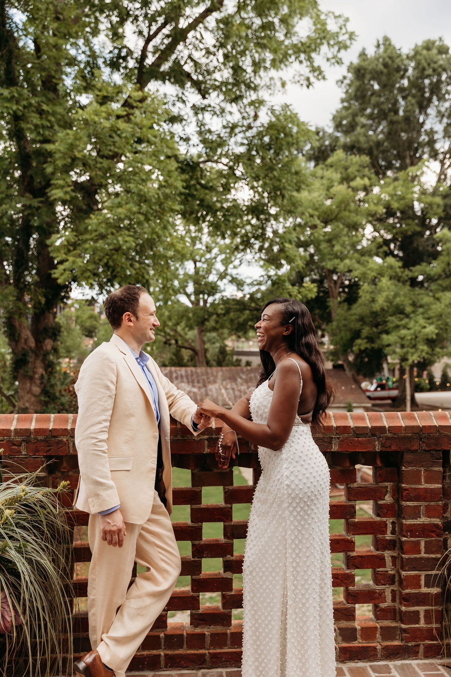 Bride and groom share a playful moment laughing together by a decorative brick wall in an outdoor garden setting.