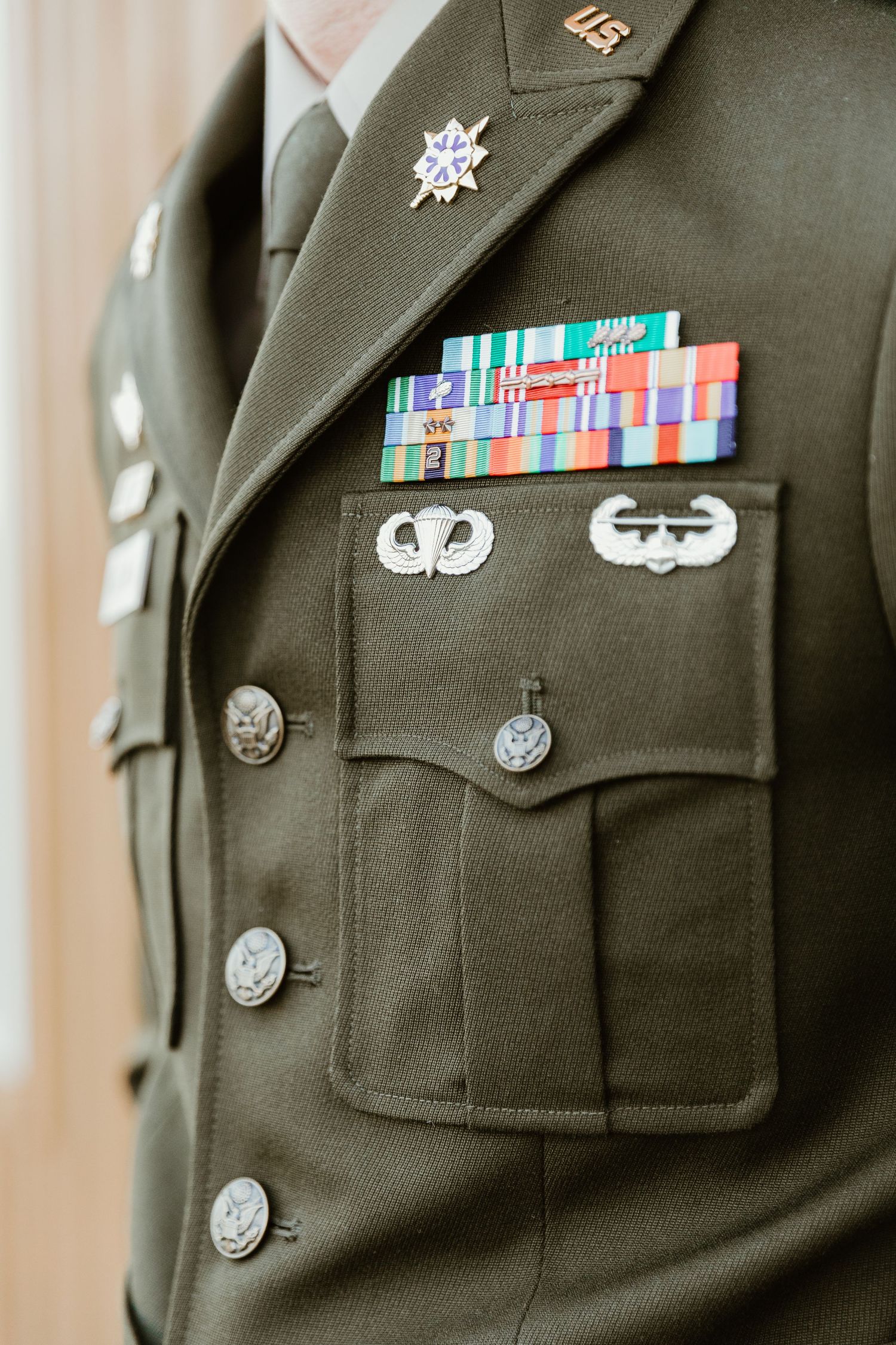 Close-up view of military dress uniform showing service ribbons, badges and rank insignia on olive green fabric.