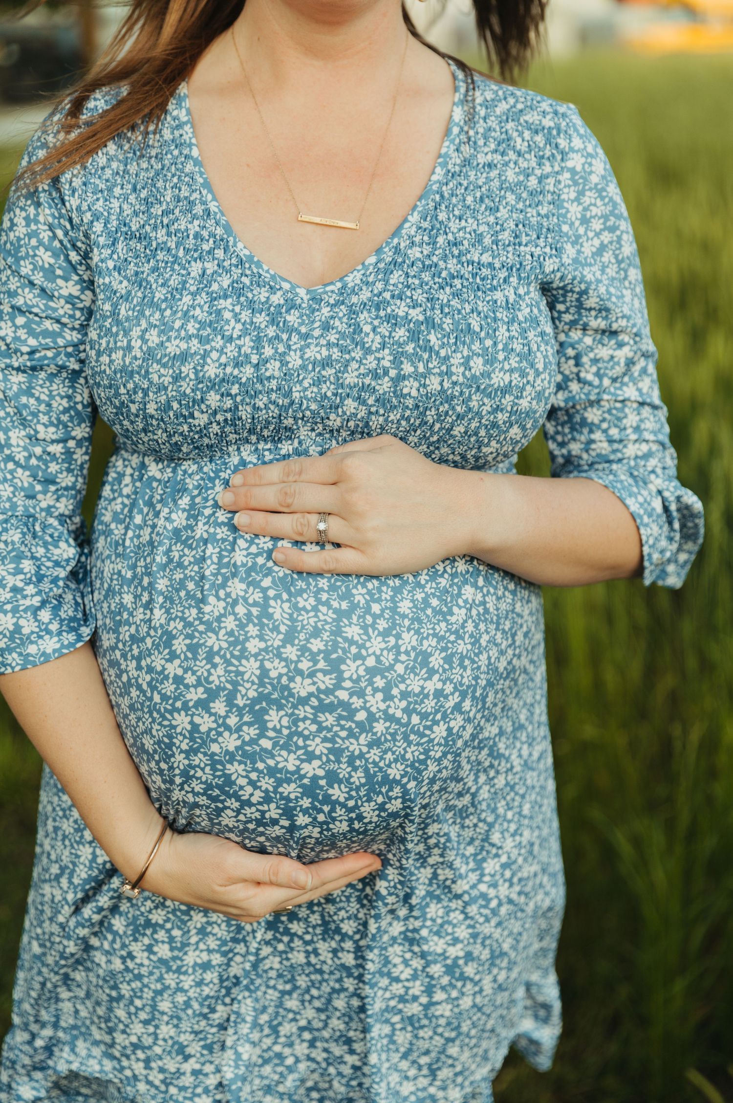 Woman in blue floral maternity dress touches her pregnant belly while standing outdoors on a North Carolina farm
