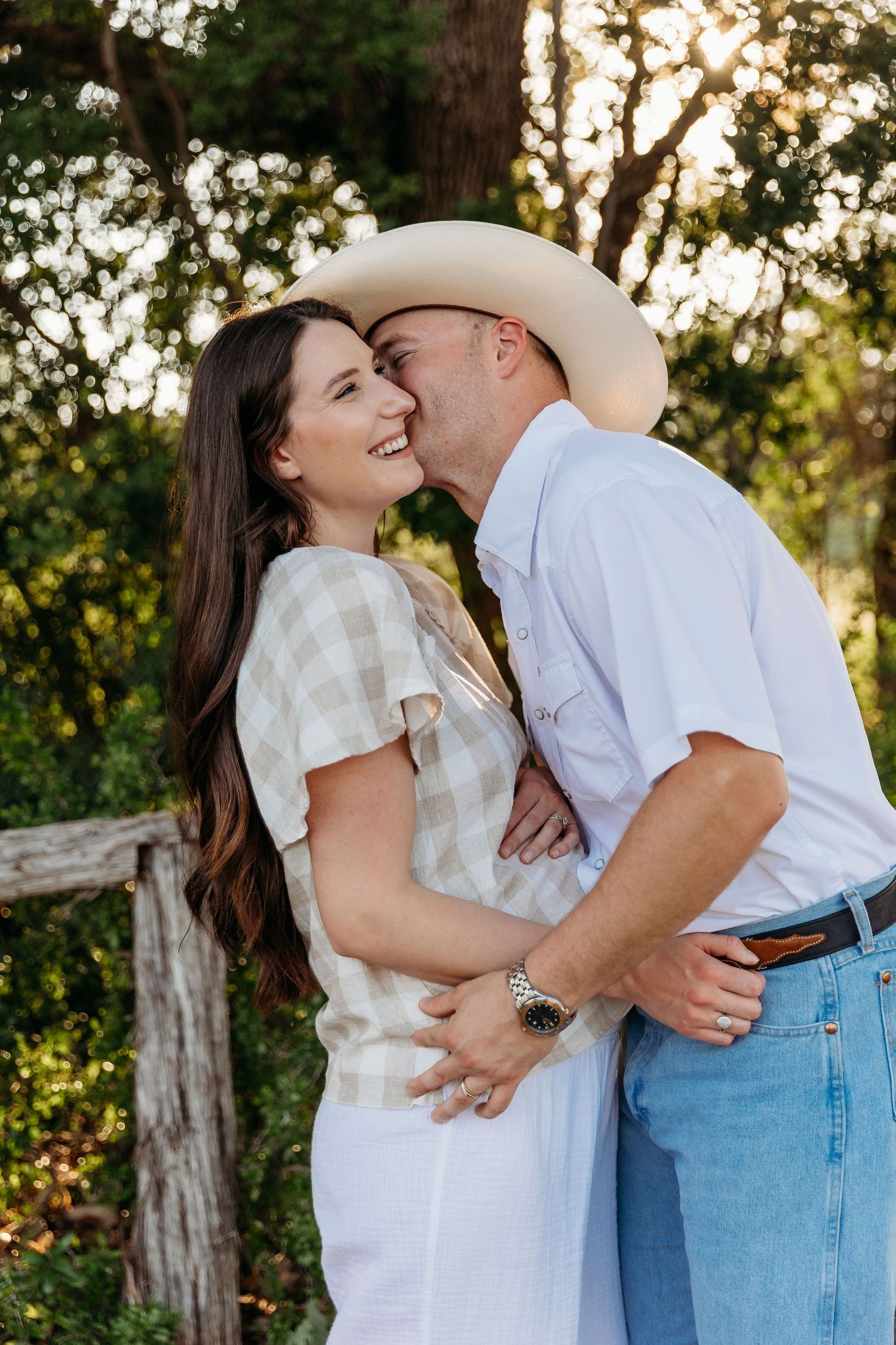 A couple in white and denim embrace and laugh together during a sunset photoshoot outdoors.