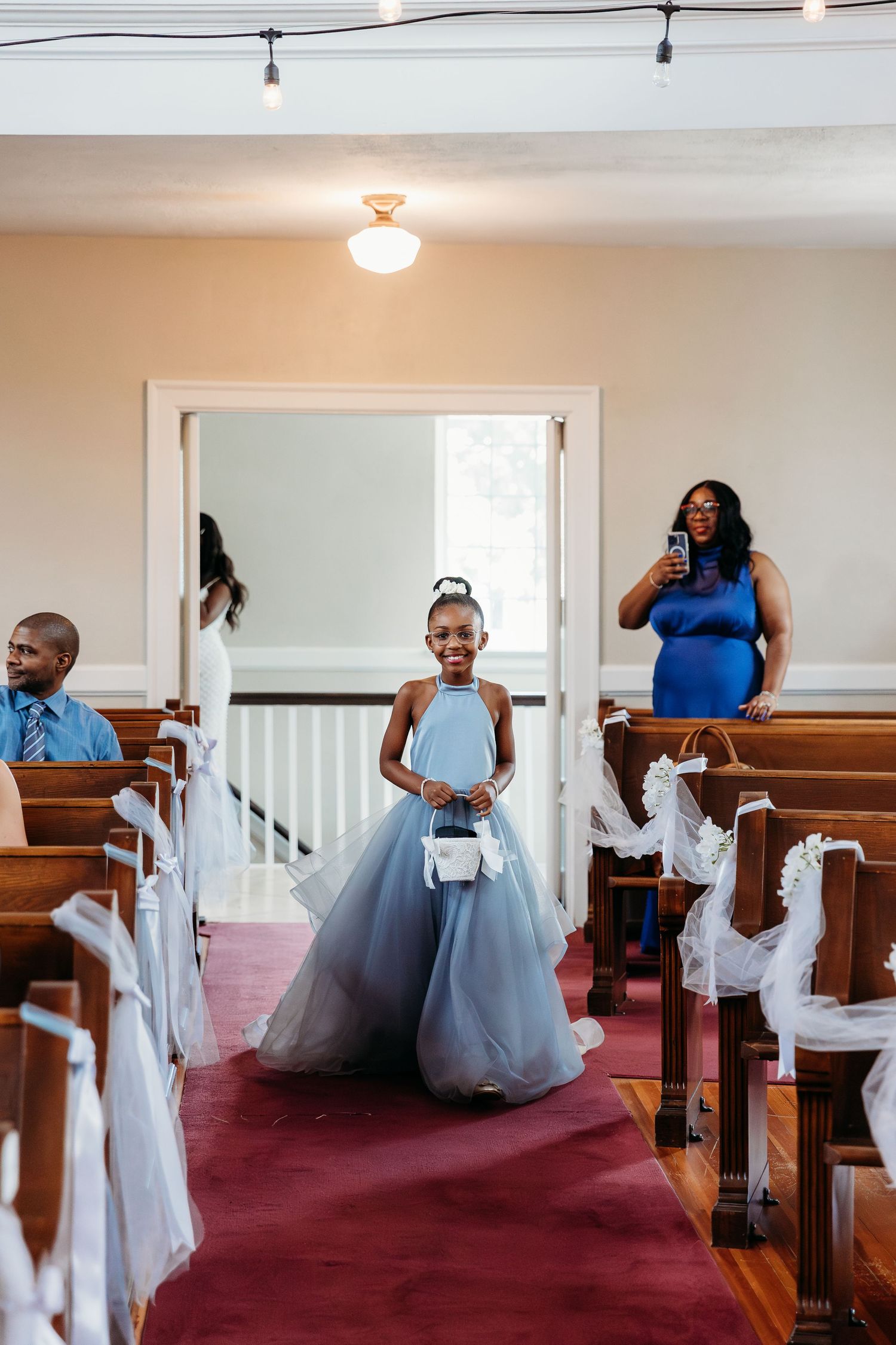 Young attendant in a light blue tulle gown walks down the aisle of a church with wooden pews and red carpet.
