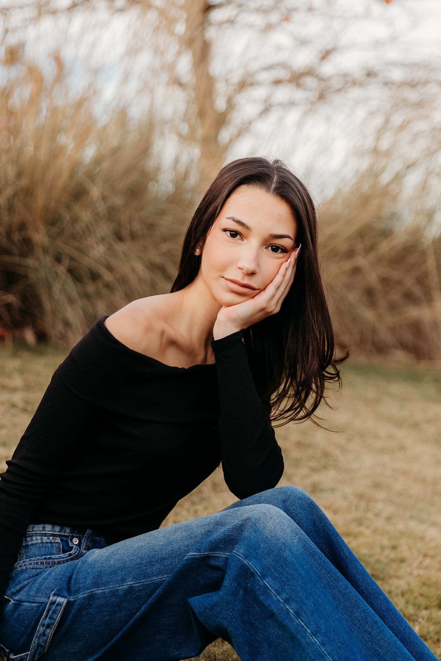 A person in a black sweater and jeans poses casually outdoors against a natural autumn background.