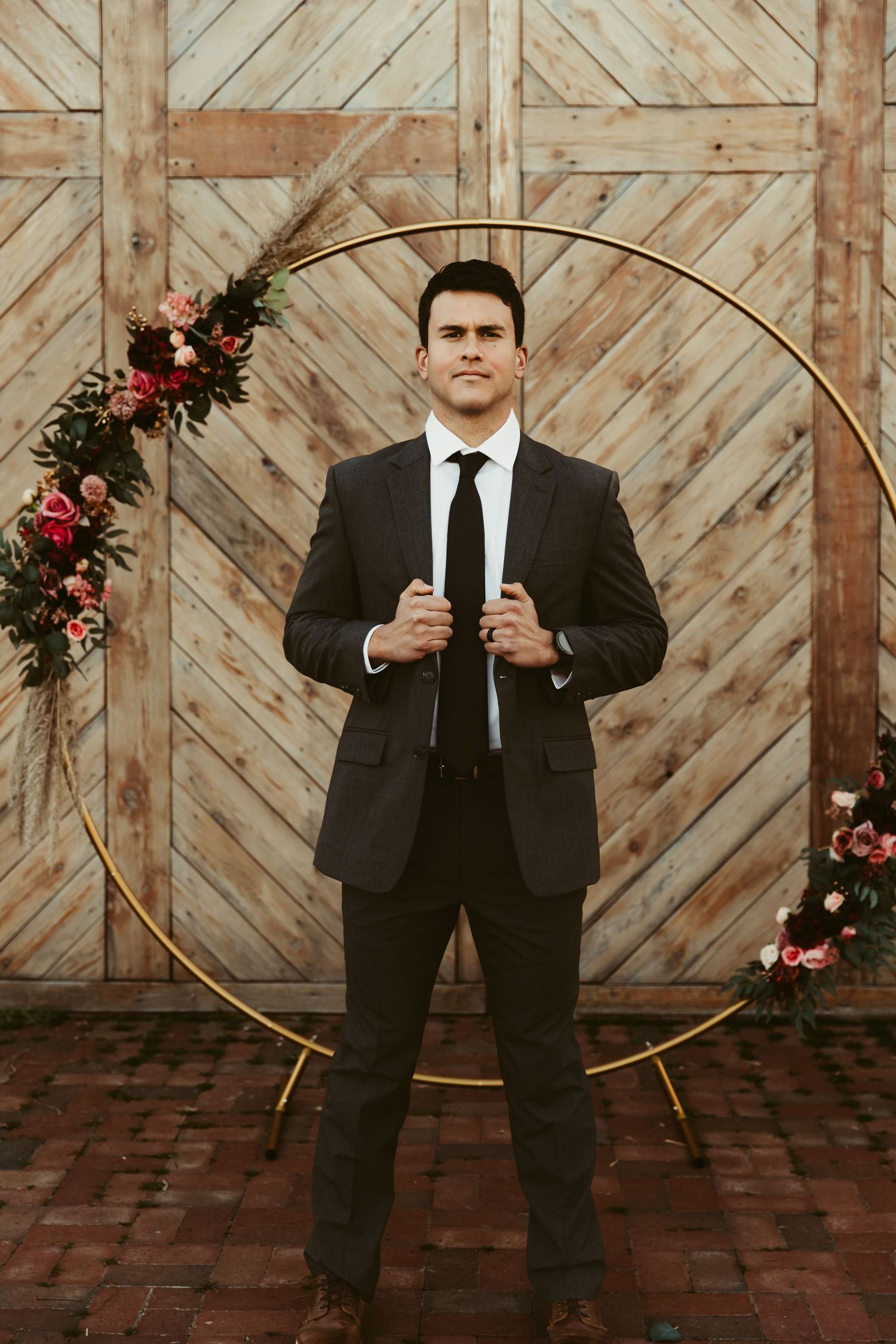 Wedding party member in black suit poses playfully against rustic wooden wall with floral decorations.