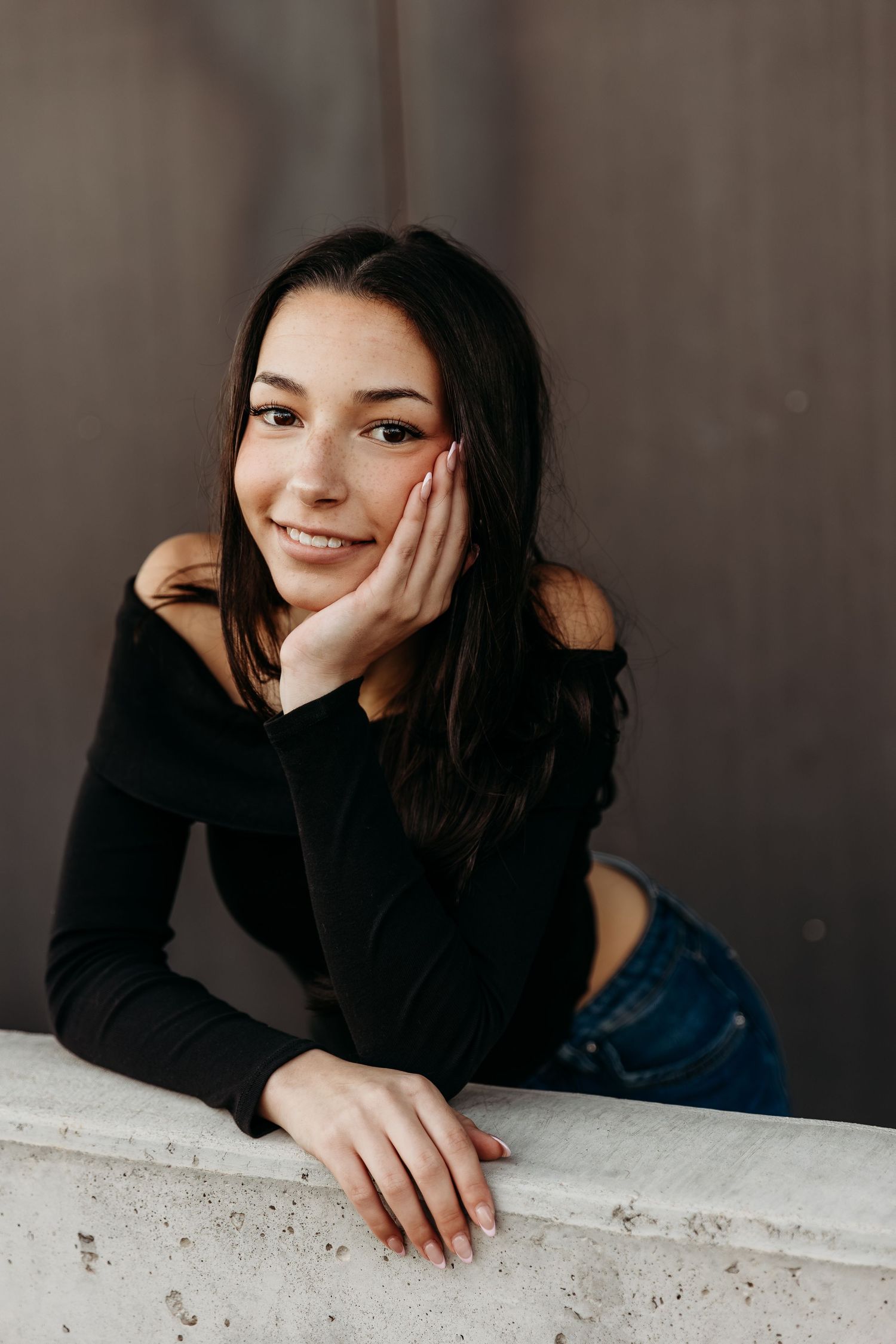 A person in a black sweater leans on a white surface while posing in a casual portrait series against a dark background.
