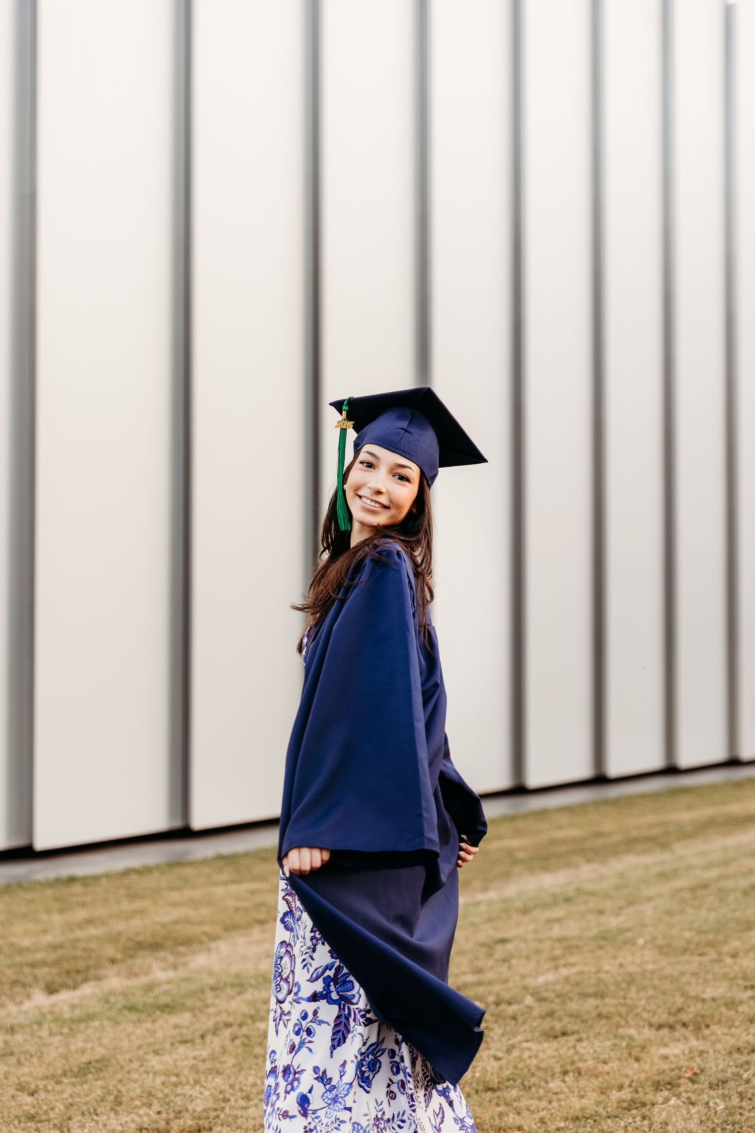 Graduate in blue cap and gown poses on grassy hill near striped wall in floral dress.