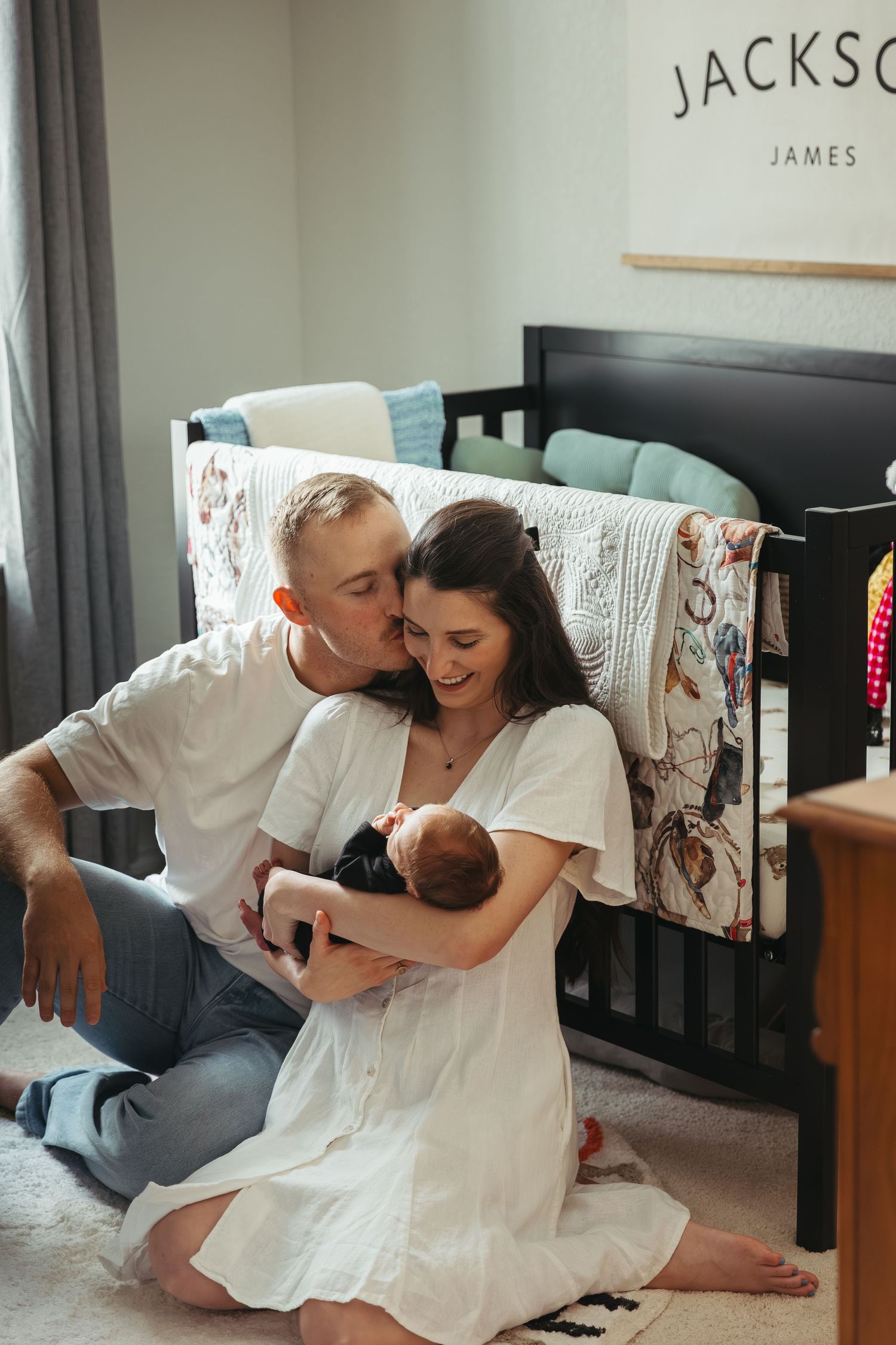 A couple in white clothing shares tender moments with their baby while sitting on the floor of a nursery.