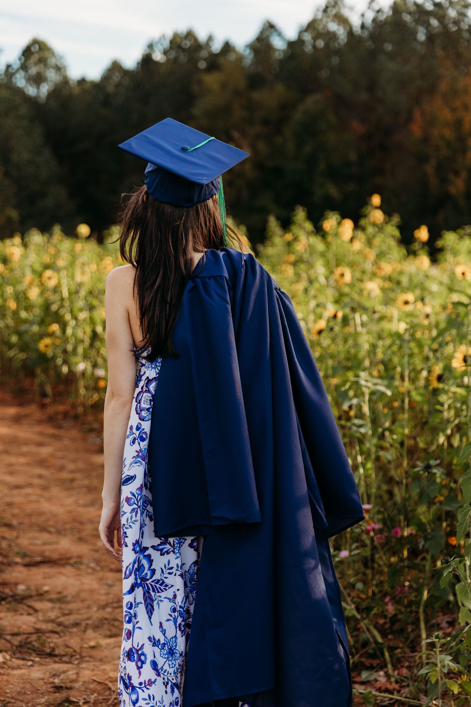 Graduate in blue and white floral dress poses in sunlit field wearing navy cap with green tassel.