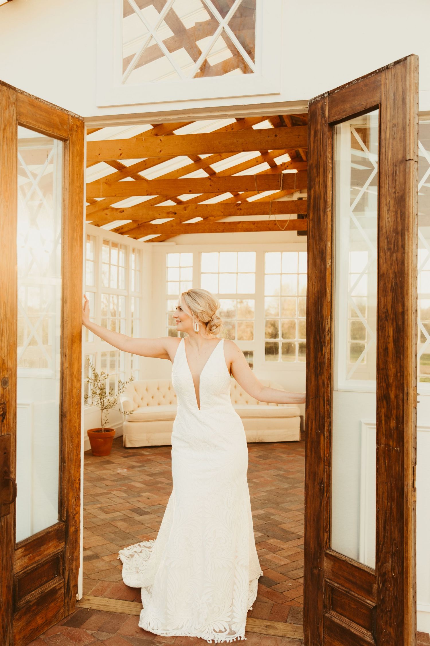 A person in a white dress stands in a rustic wooden doorway with large windows and natural light streaming in.