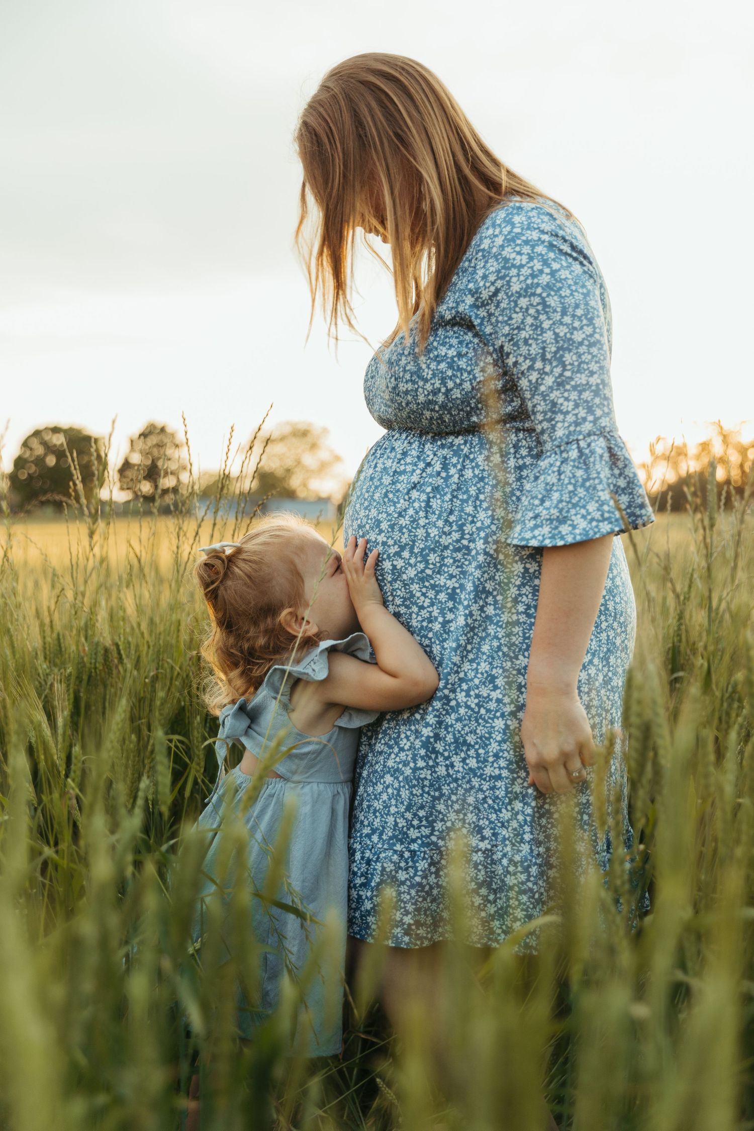 A tender moment captured in a golden wheat field as figures in blue floral dress and denim embrace at sunset.
