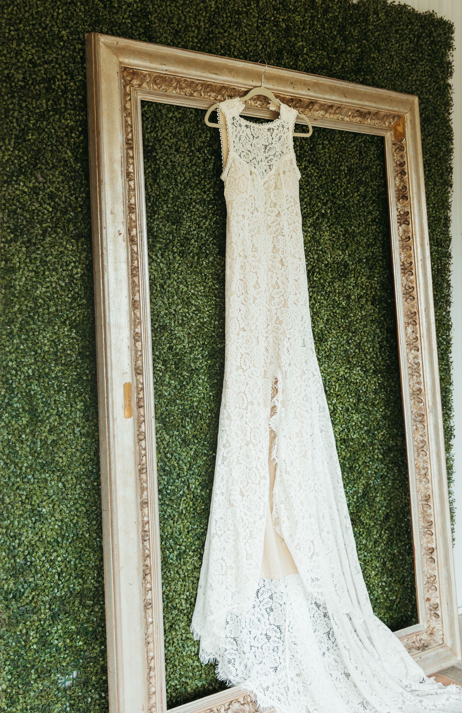 Elegant white lace wedding dress hangs on display against green hedge wall with ornate vintage frame backdrop.