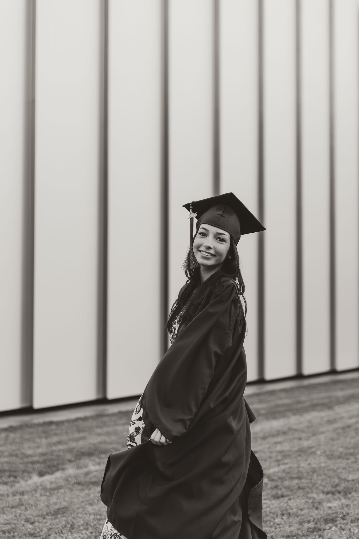 A graduate in blue cap and gown poses outdoors against a striped wall background in black and white photography.