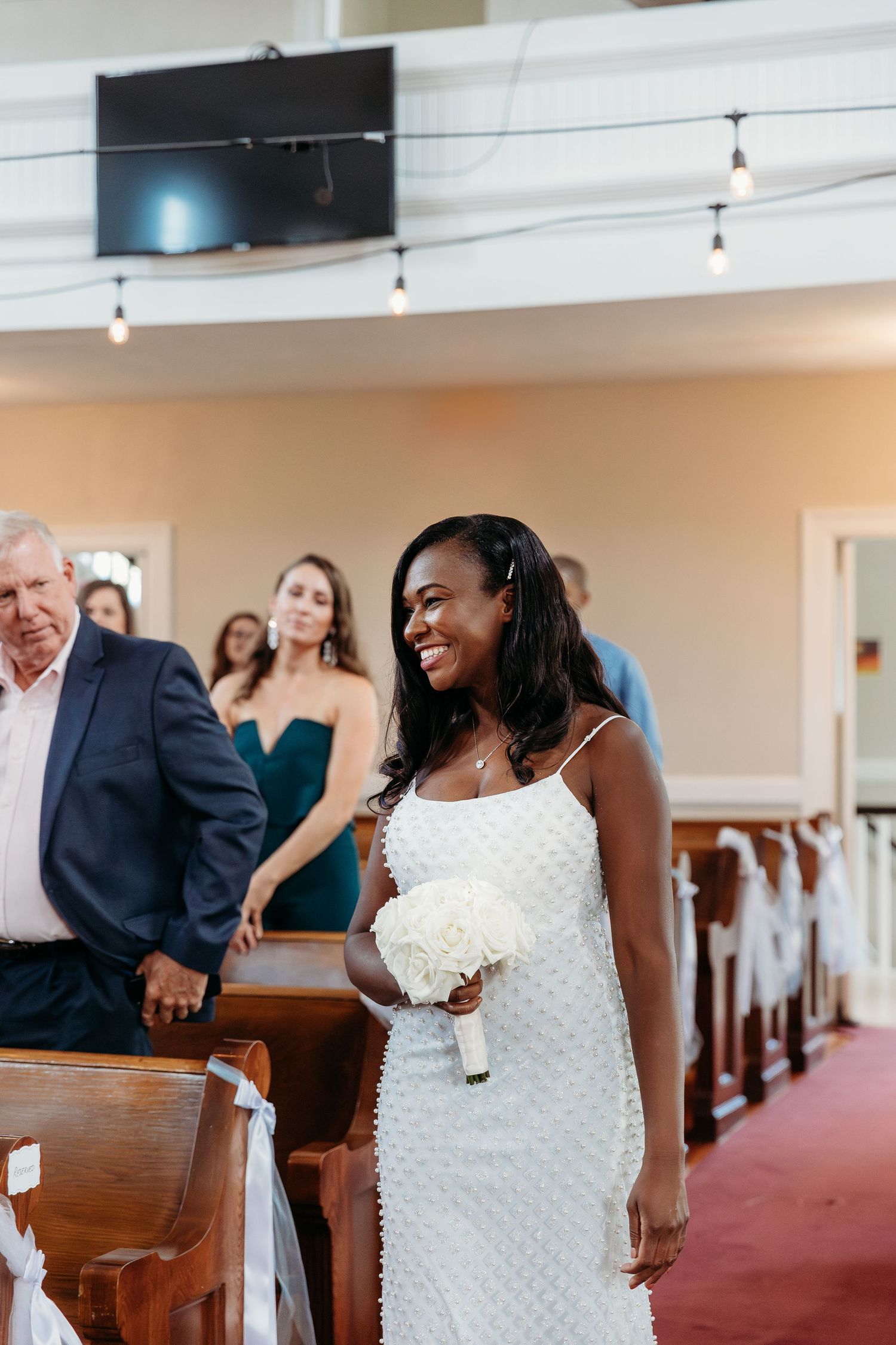 A bride in a white beaded dress stands smiling in a church aisle during her wedding ceremony.