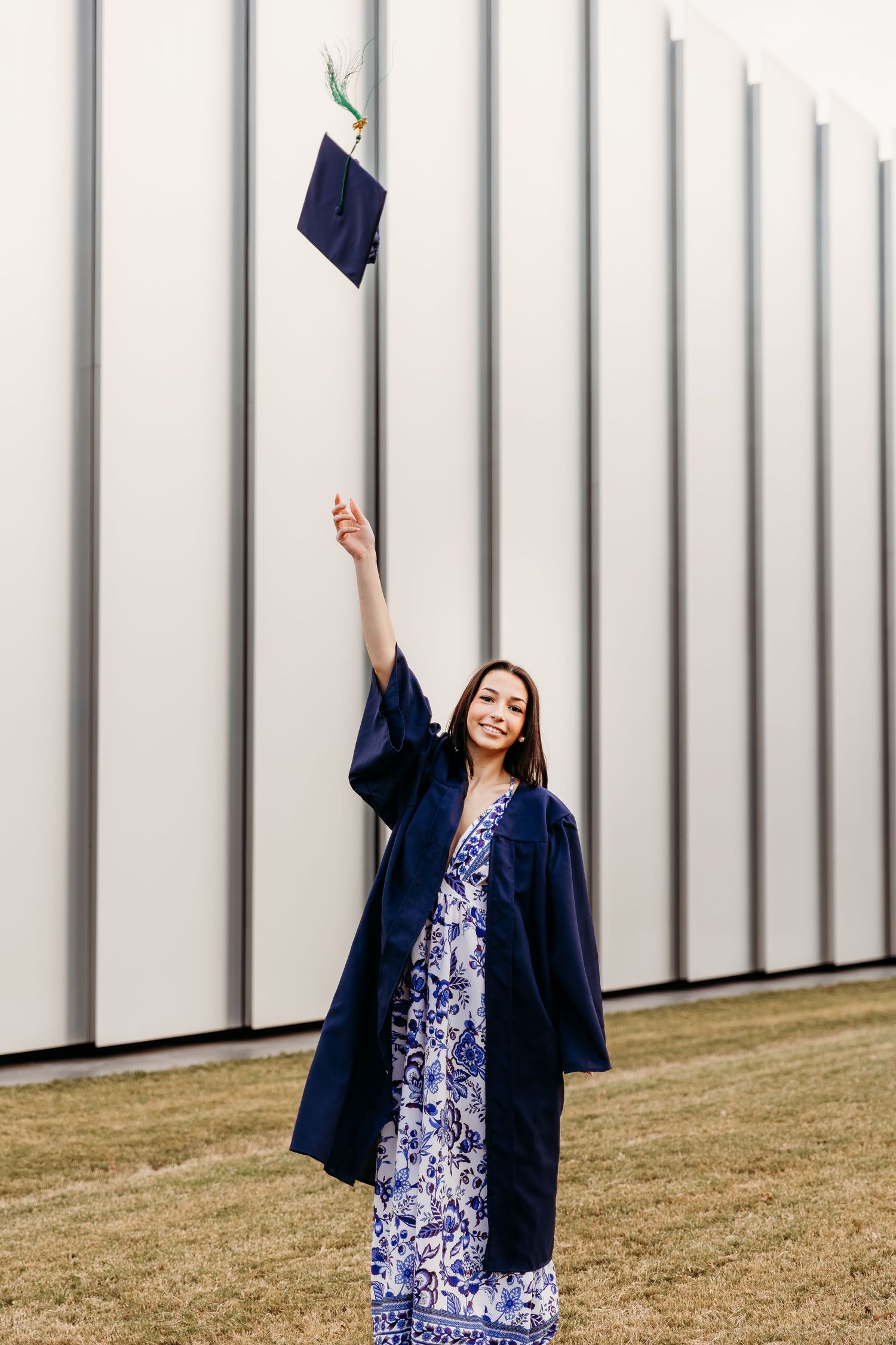 A graduate in a blue floral dress and navy gown tosses their cap against a white wall during a celebration photoshoot.