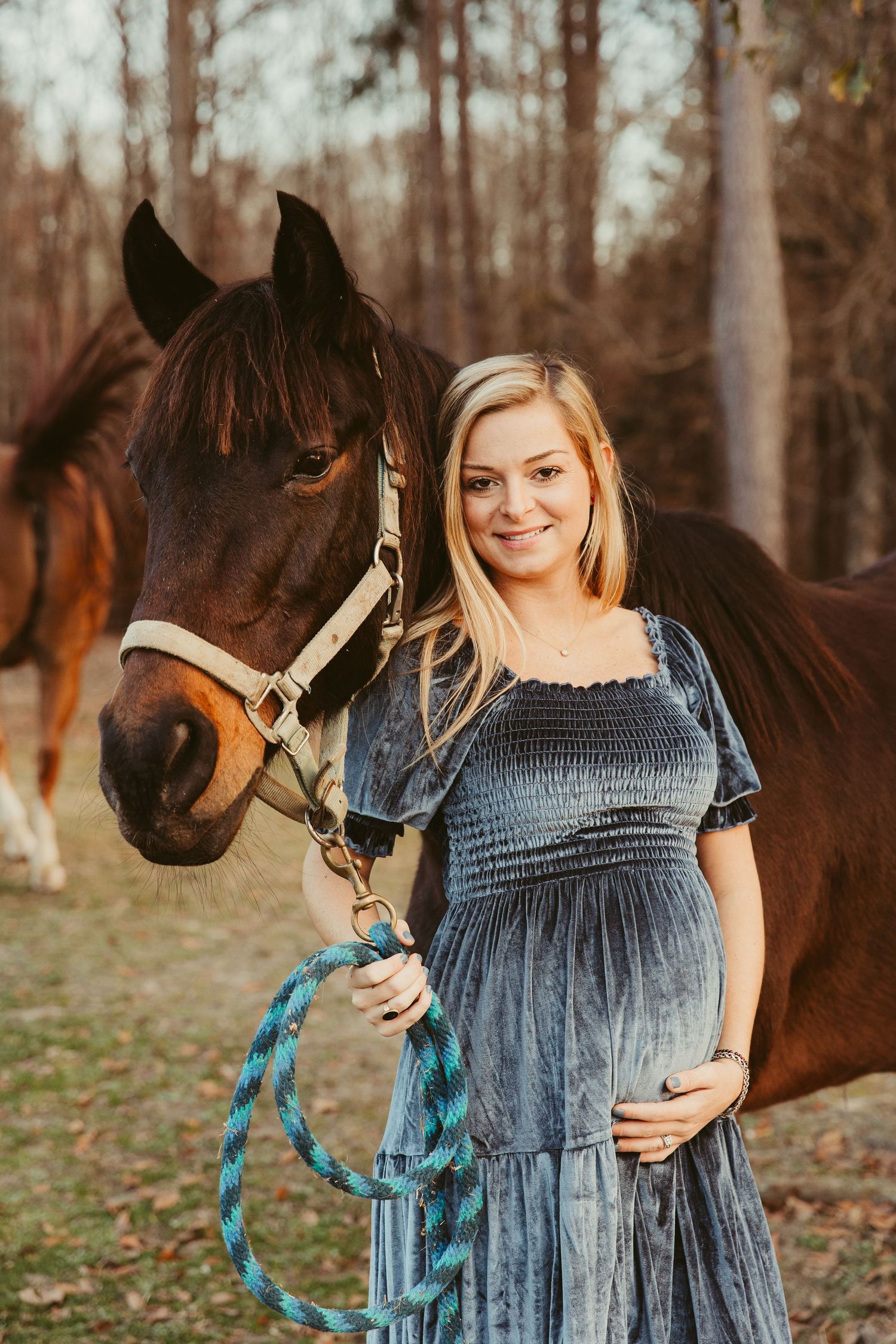A woman in a blue dress stands beside a brown horse in an autumn forest setting.