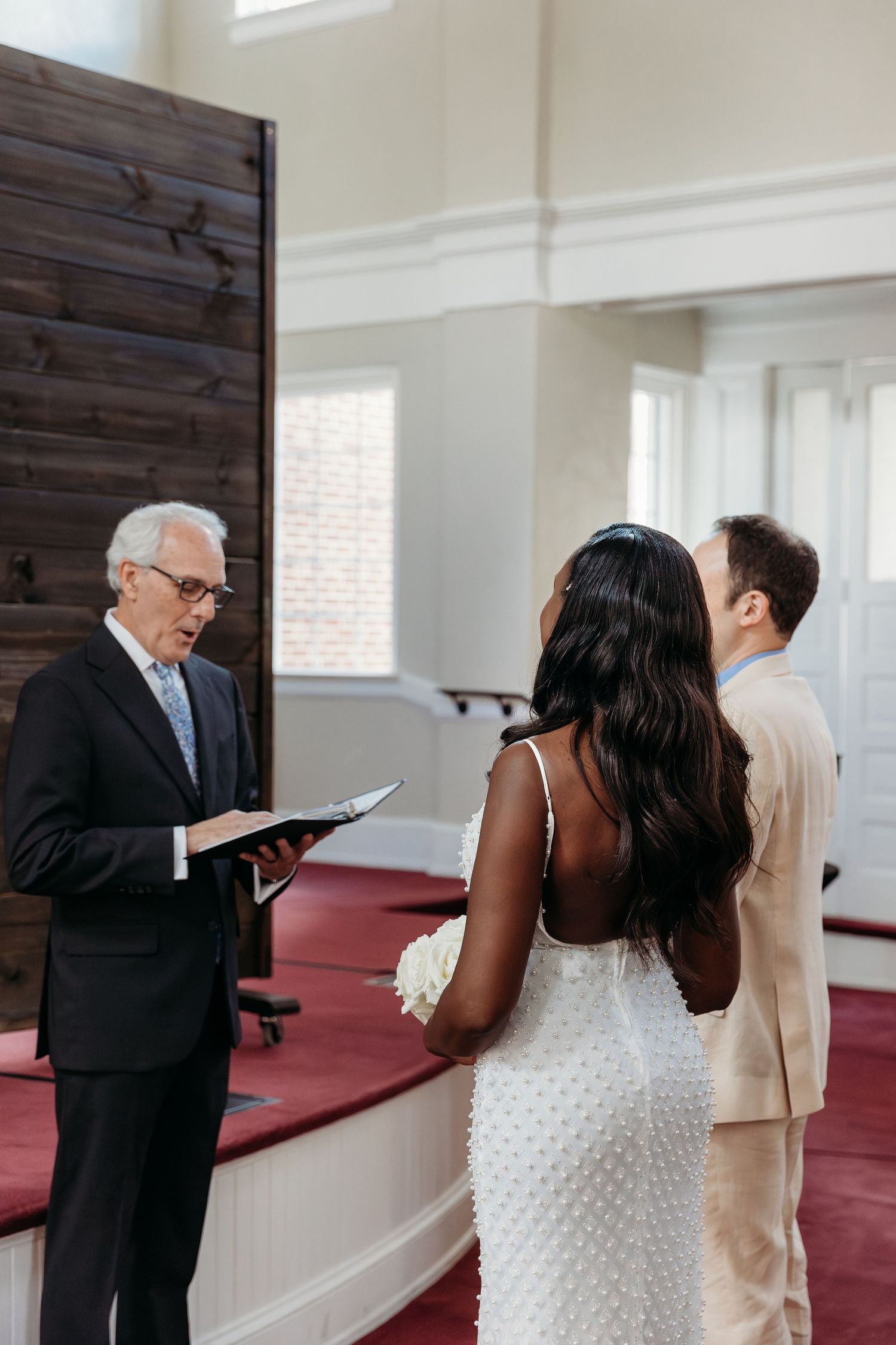 A couple in formal attire stands together in a church with white walls and red carpeting during a ceremony.