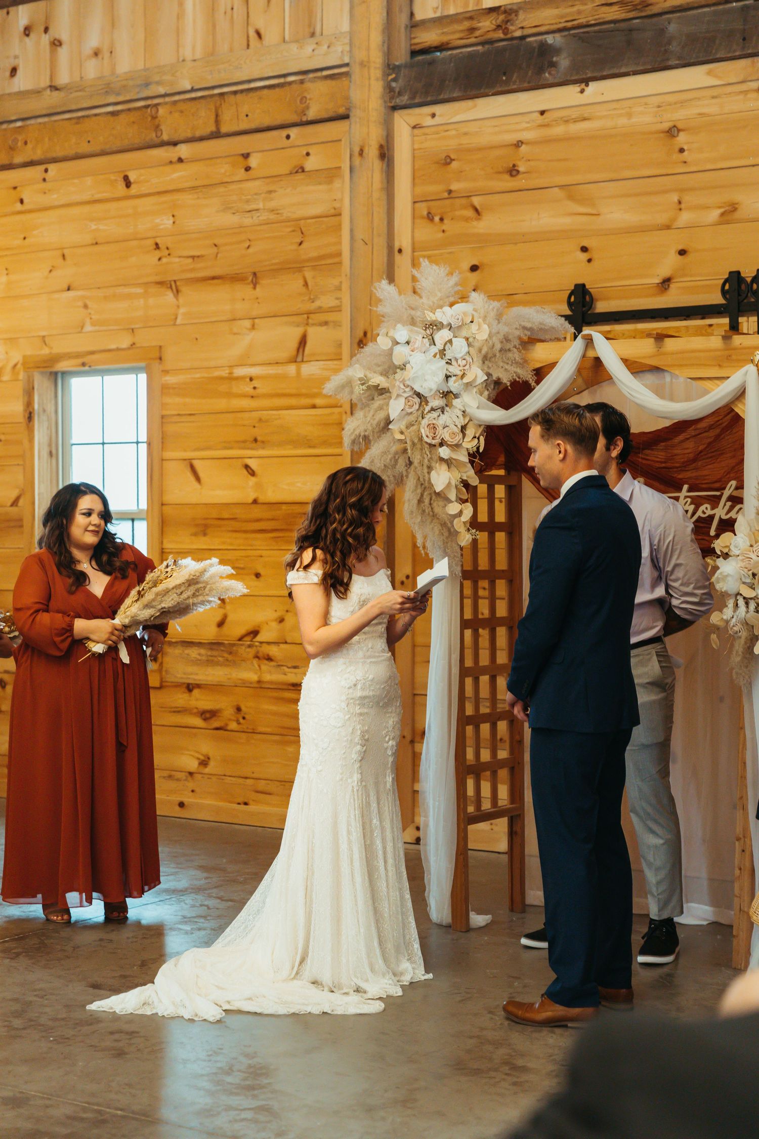 Wedding party gathers in a rustic wooden barn venue decorated with white drapes and natural light from windows.