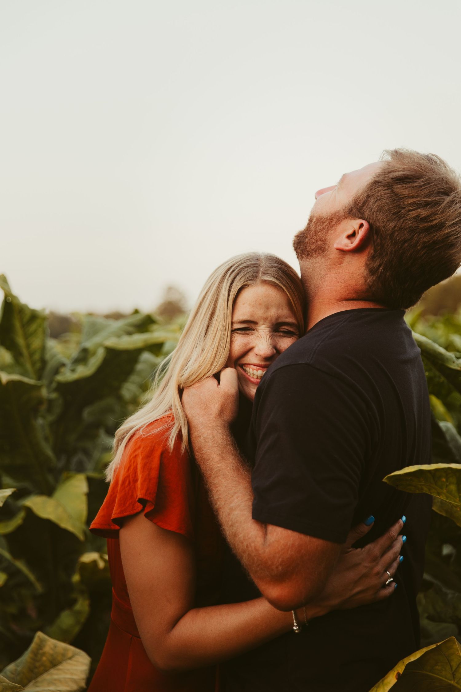 A couple embraces intimately in a natural outdoor setting with green foliage in the background at sunset.