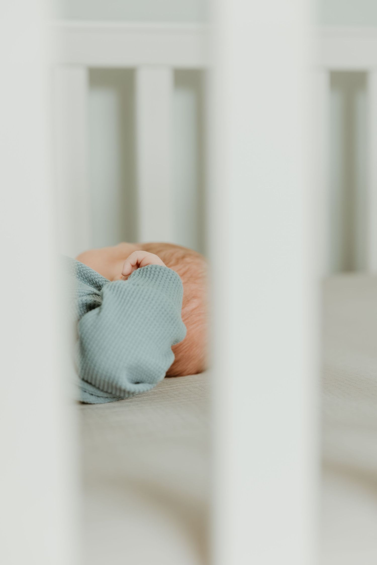 View through white crib slats showing a sleeping baby in a soft mint green outfit.