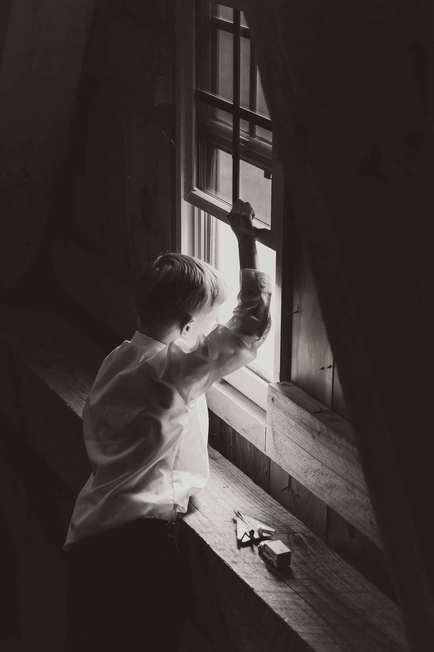 A child reaches up towards a window in a dramatic black and white photograph with soft natural light streaming in.