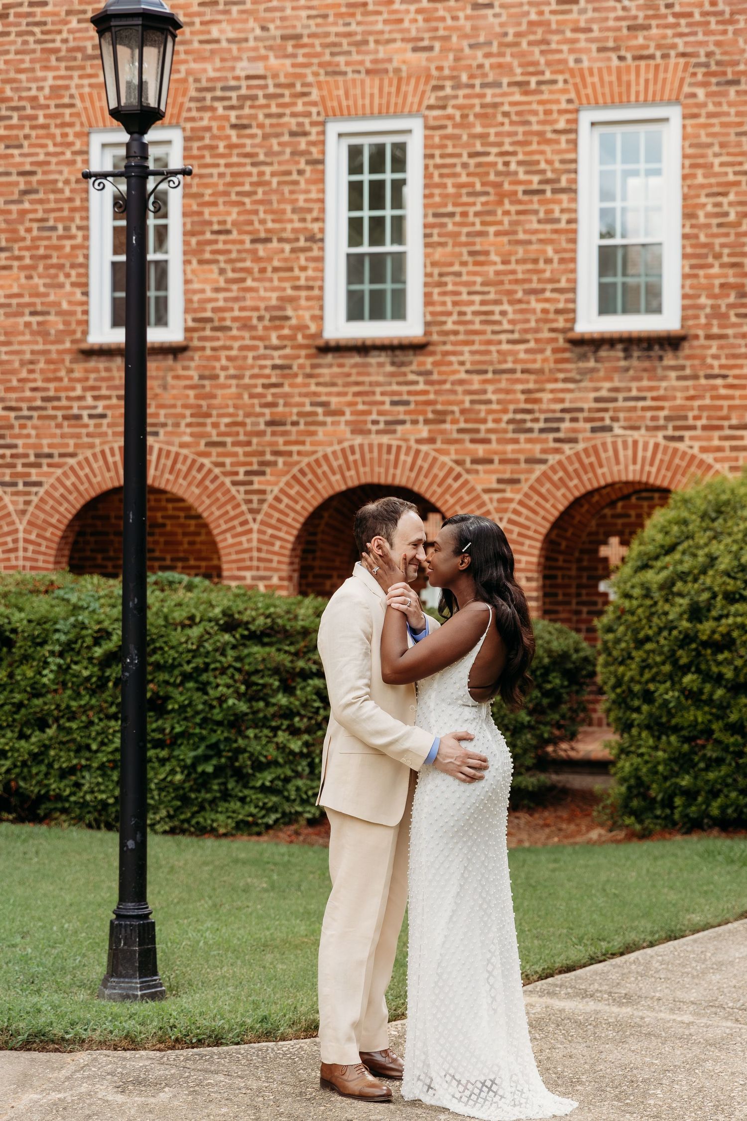 A romantic couple shares a tender moment in front of a historic brick building with arched windows and lamp posts.