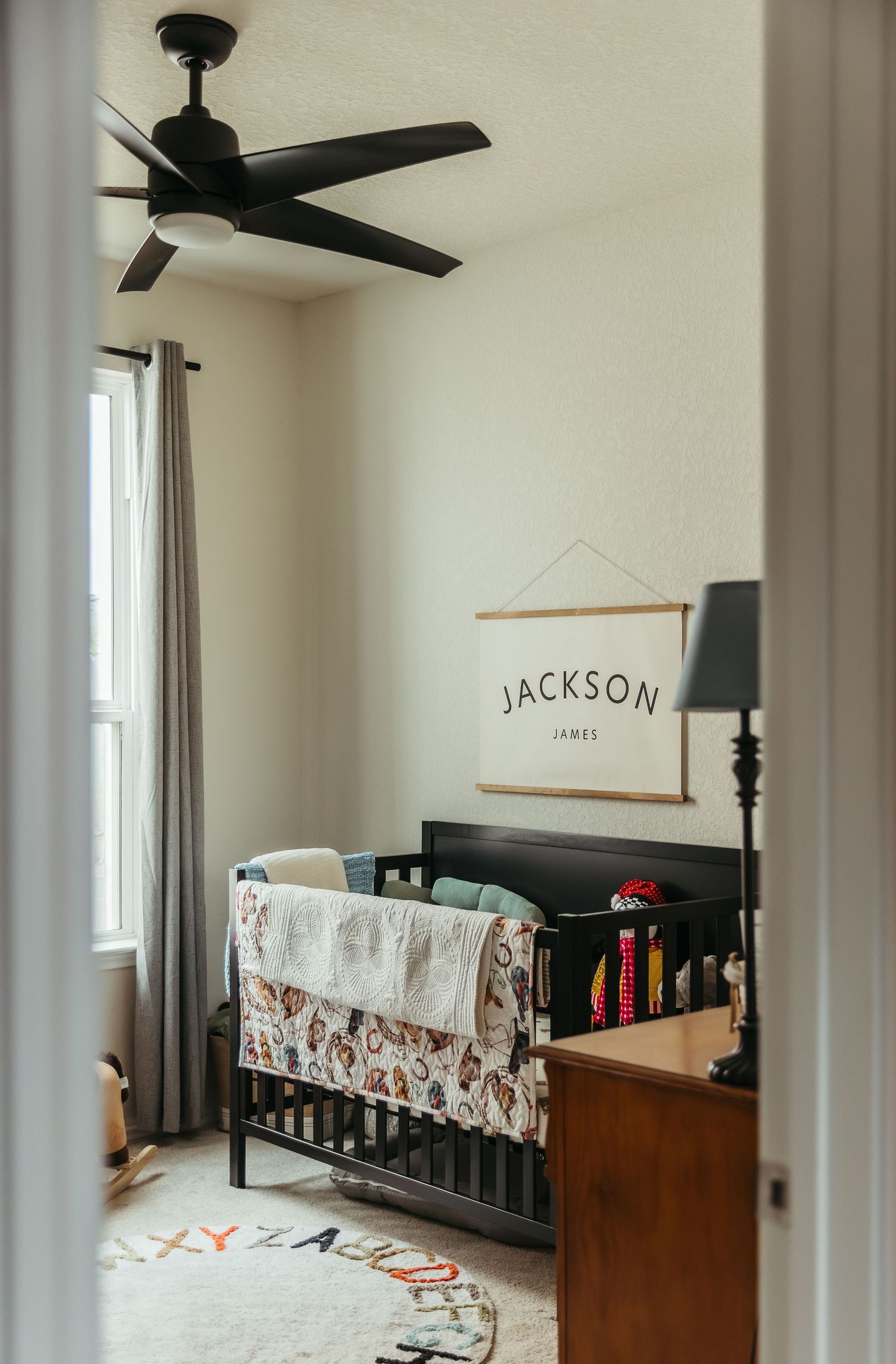 Modern nursery room with dark wood crib, ceiling fan, and white walls featuring 'Jackson' wall decor.