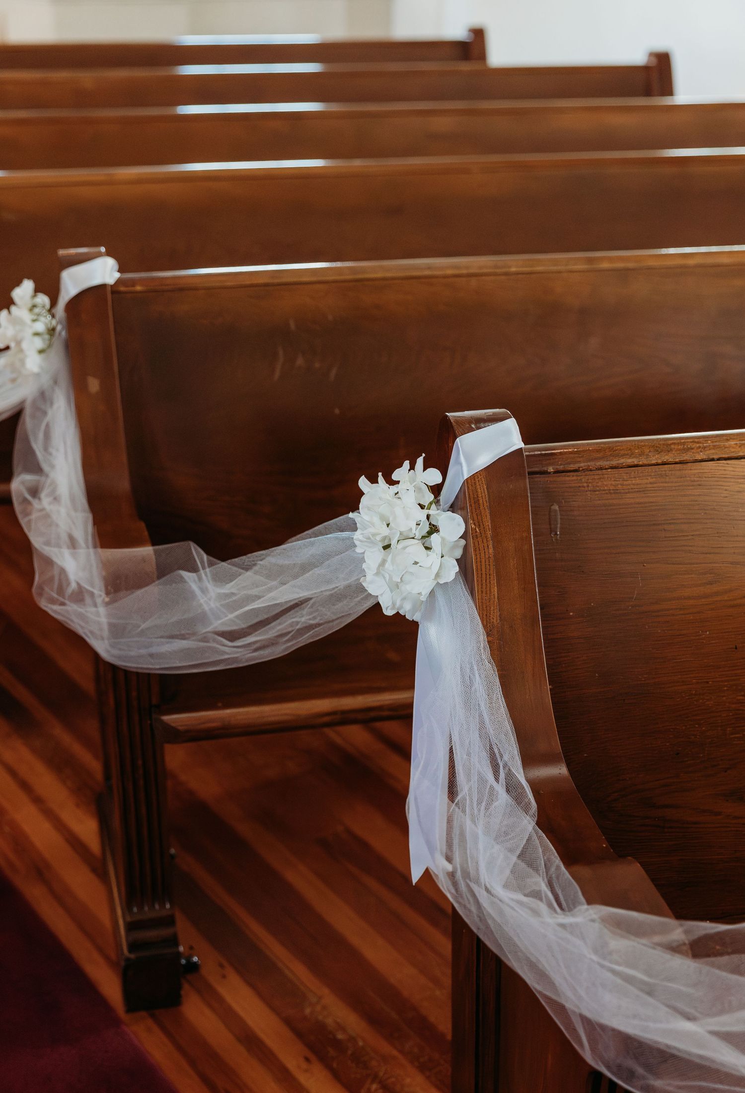 Church pews decorated with delicate white tulle and floral arrangements for a wedding ceremony.